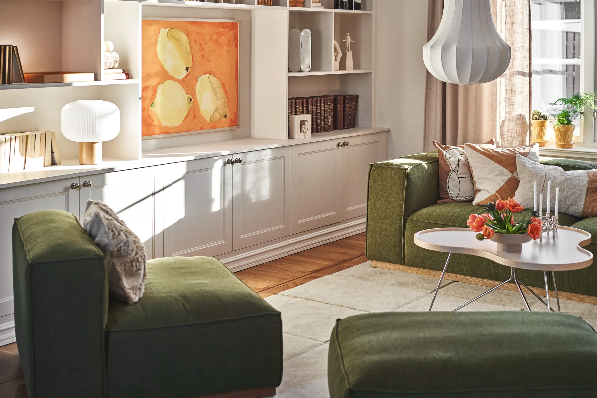 Zonnige woonkamer met groene banken, witte salontafel met oranje tulpen, kaarsen en witte ingebouwde planken met kunst.