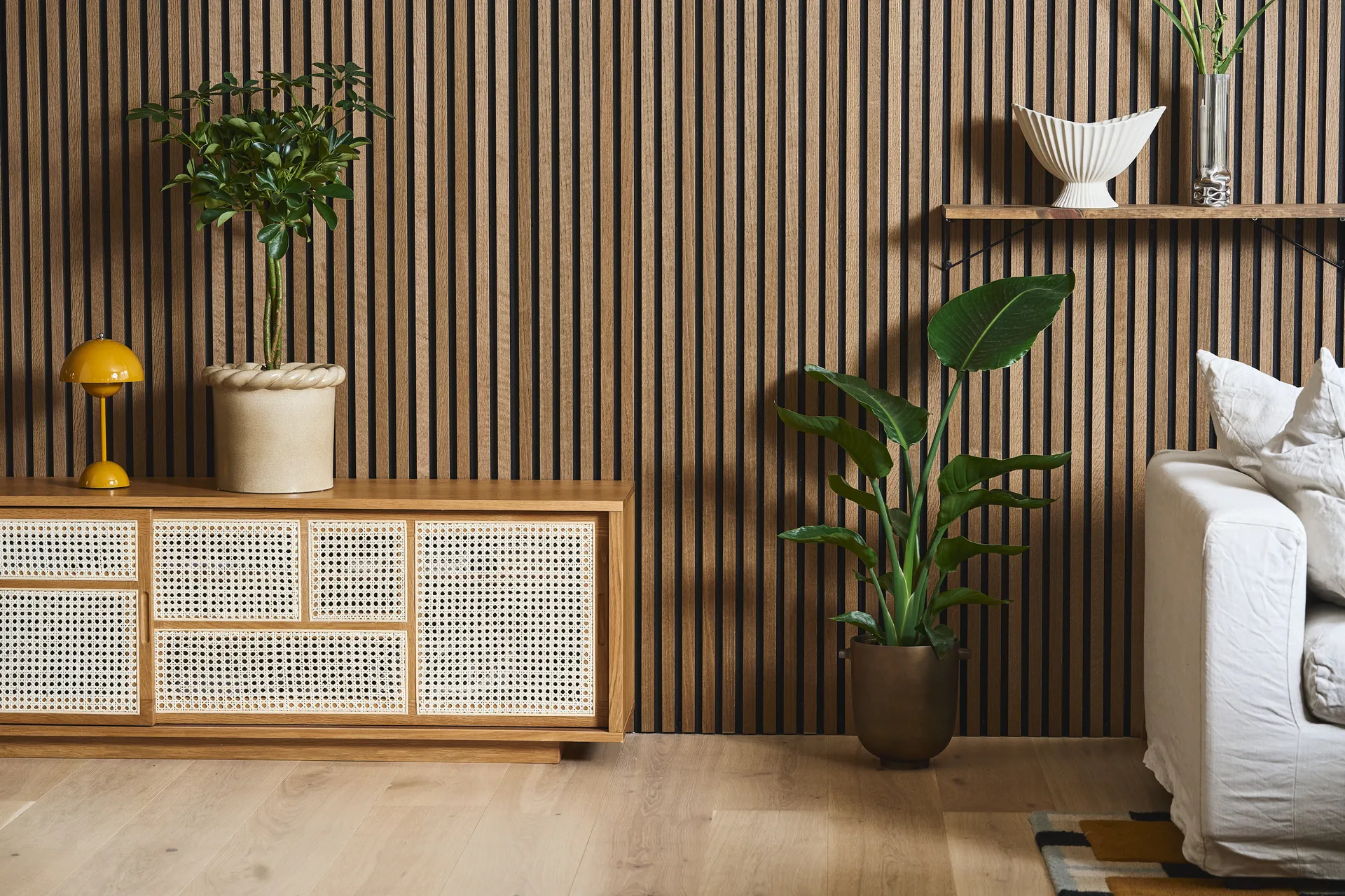 Moderne woonkamer met lattenwand, rotan kast, gele lamp, diverse potplanten en een witte bank.