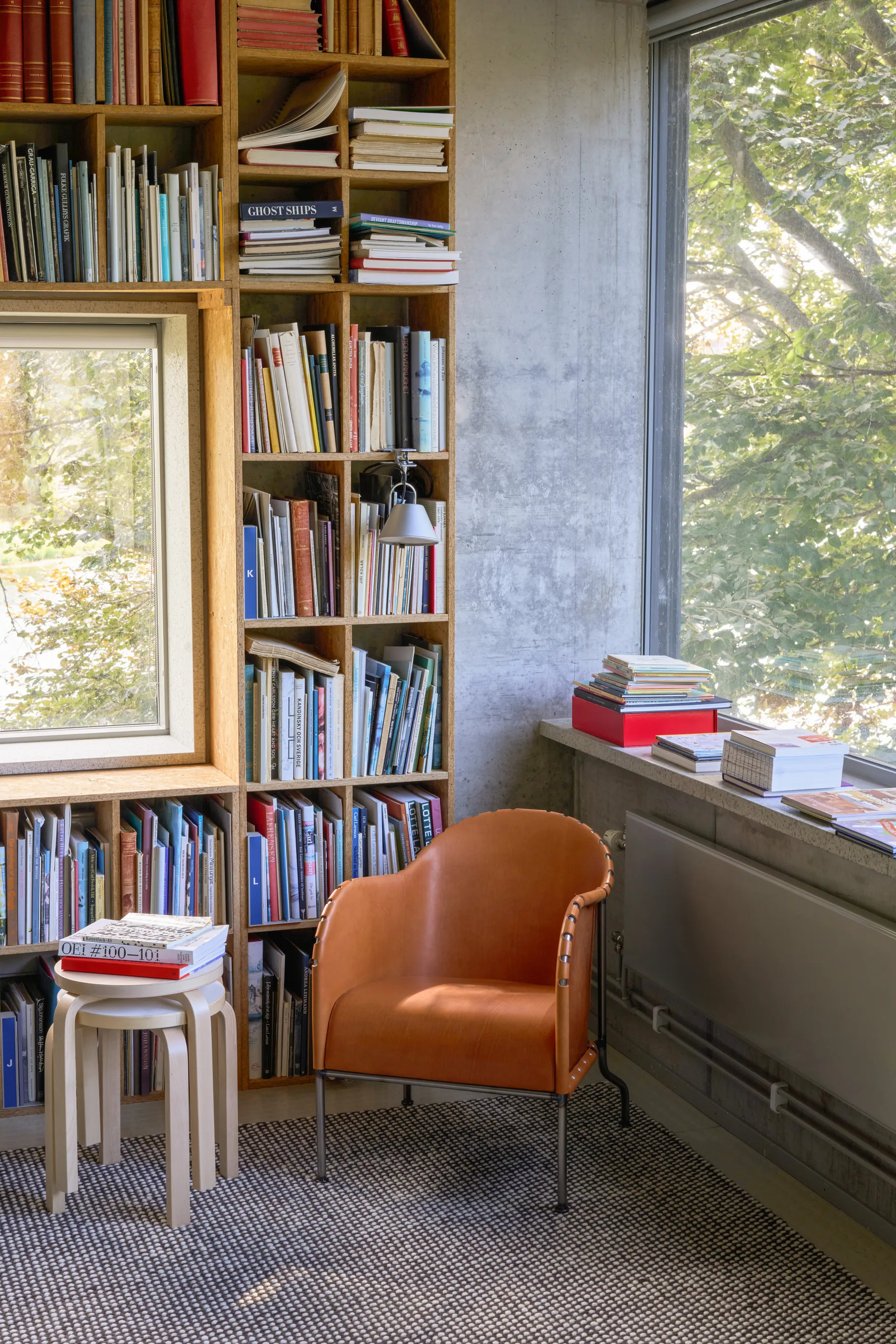 Gezellige kamer met grote houten boekenkast, lichtbruine leren fauteuil, kleine kruk met boeken en raam met uitzicht op bomen.