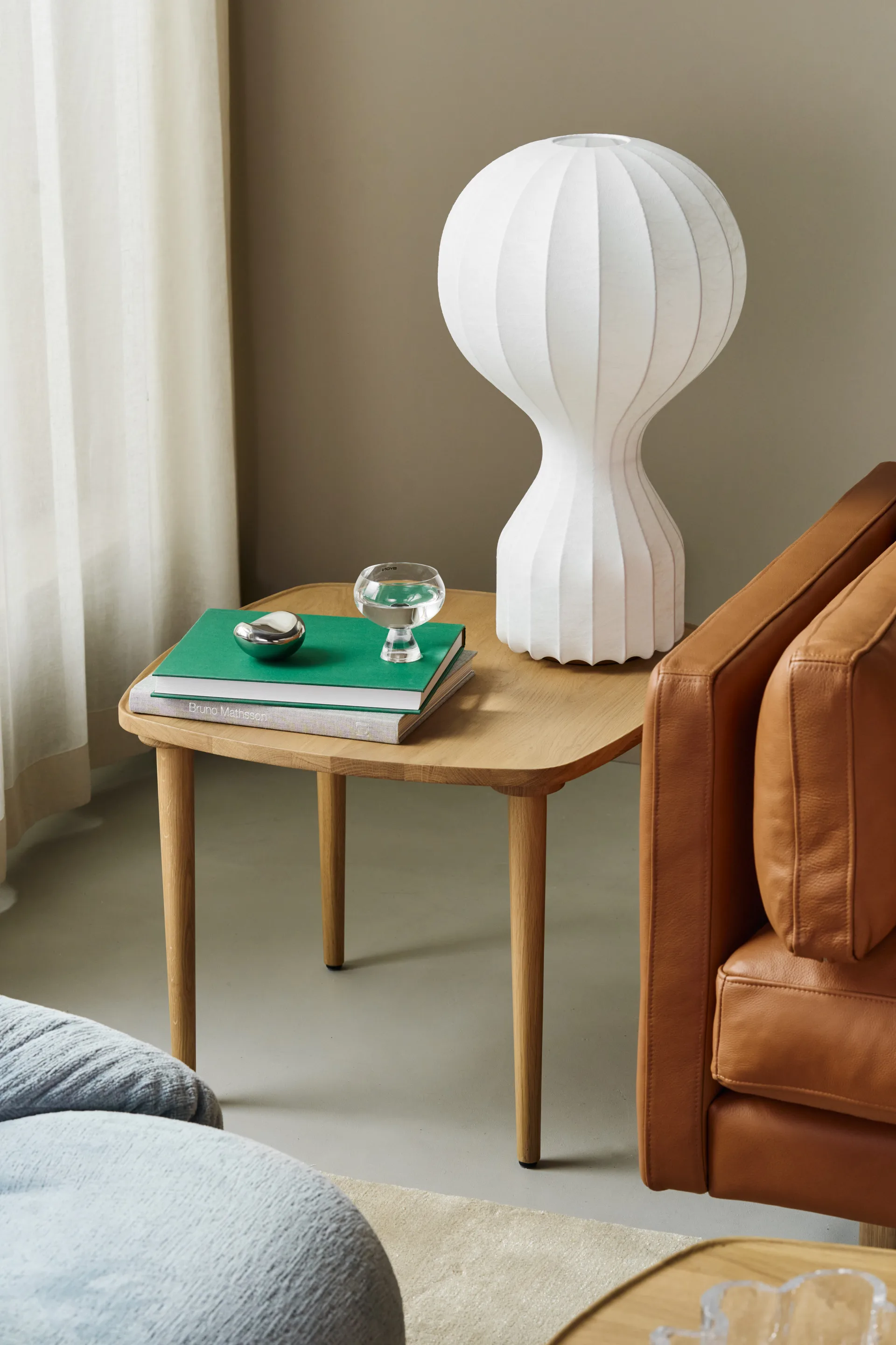 Lichte houten bijzettafel met witte geribbelde lamp, groene boeken, een glas water en een zilveren object, naast bruine leren stoel.