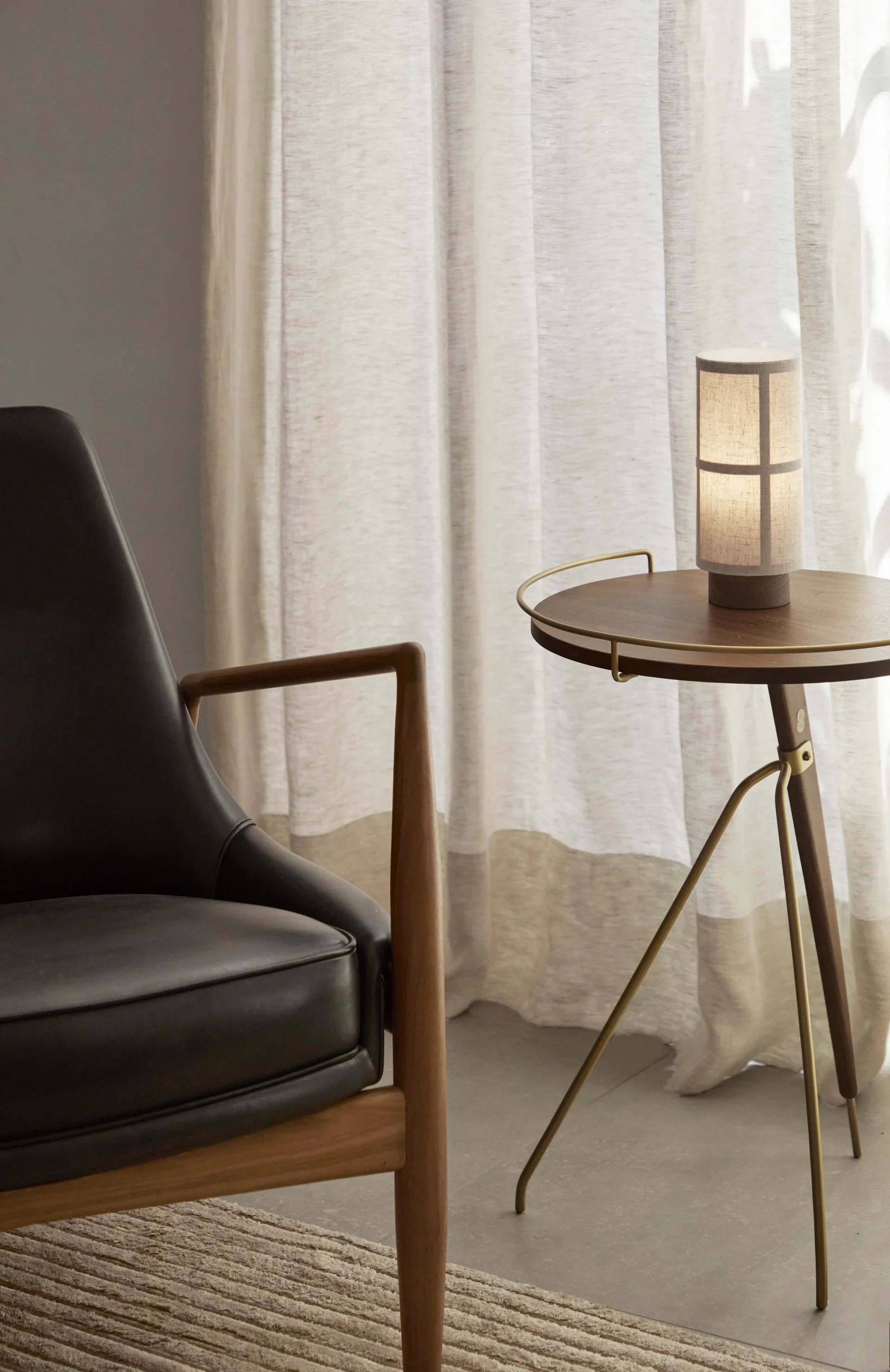 Audo Copenhagen zwarte leren fauteuil en ronde houten bijzettafel met brandende lamp. Lichte gordijnen.