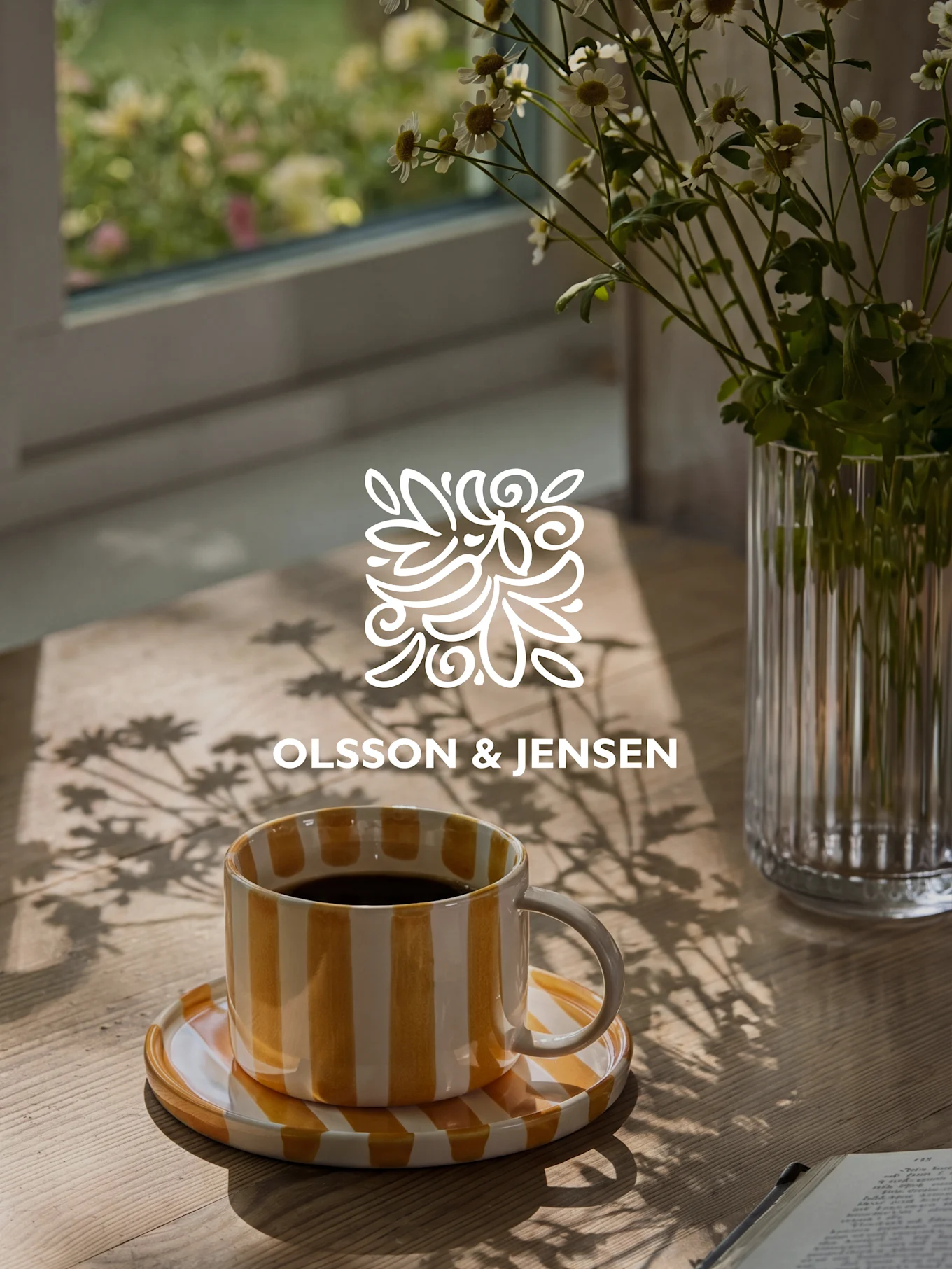 Gele en wit gestreepte koffiemok op een houten tafel met een vaas witte bloemen en zonlichtschaduwen, Olsson & Jensen logo.