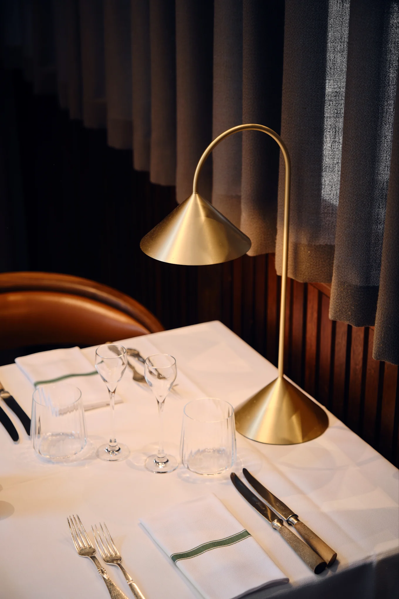 Een moderne gouden tafellamp verlicht een gedekte restauranttafel met wit linnen, glazen, bestek en een servet met een groene streep.