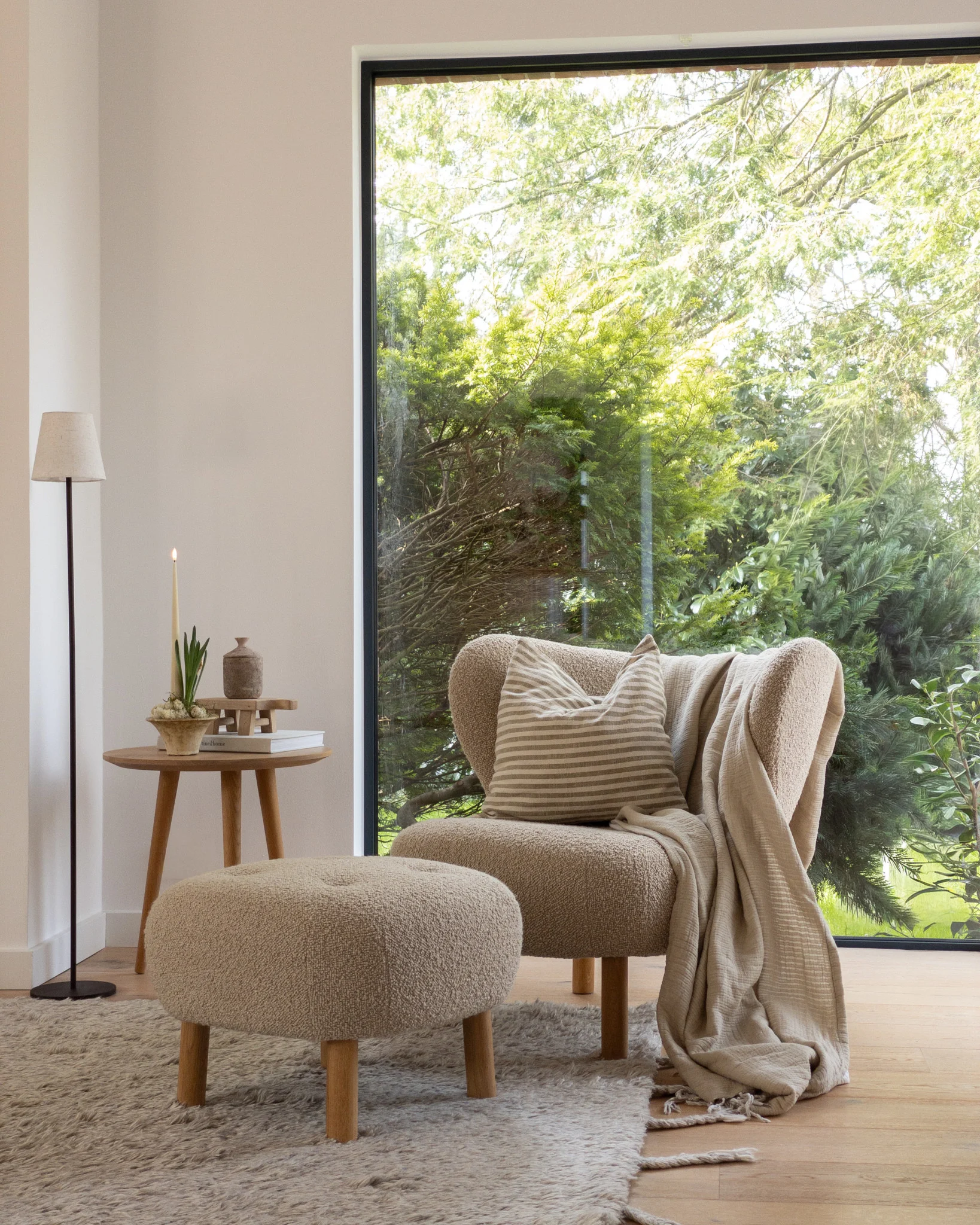 Gezellige hoek: lichtbruine fauteuil, poef, bijzettafel met kaars, groot raam, groene bomen.