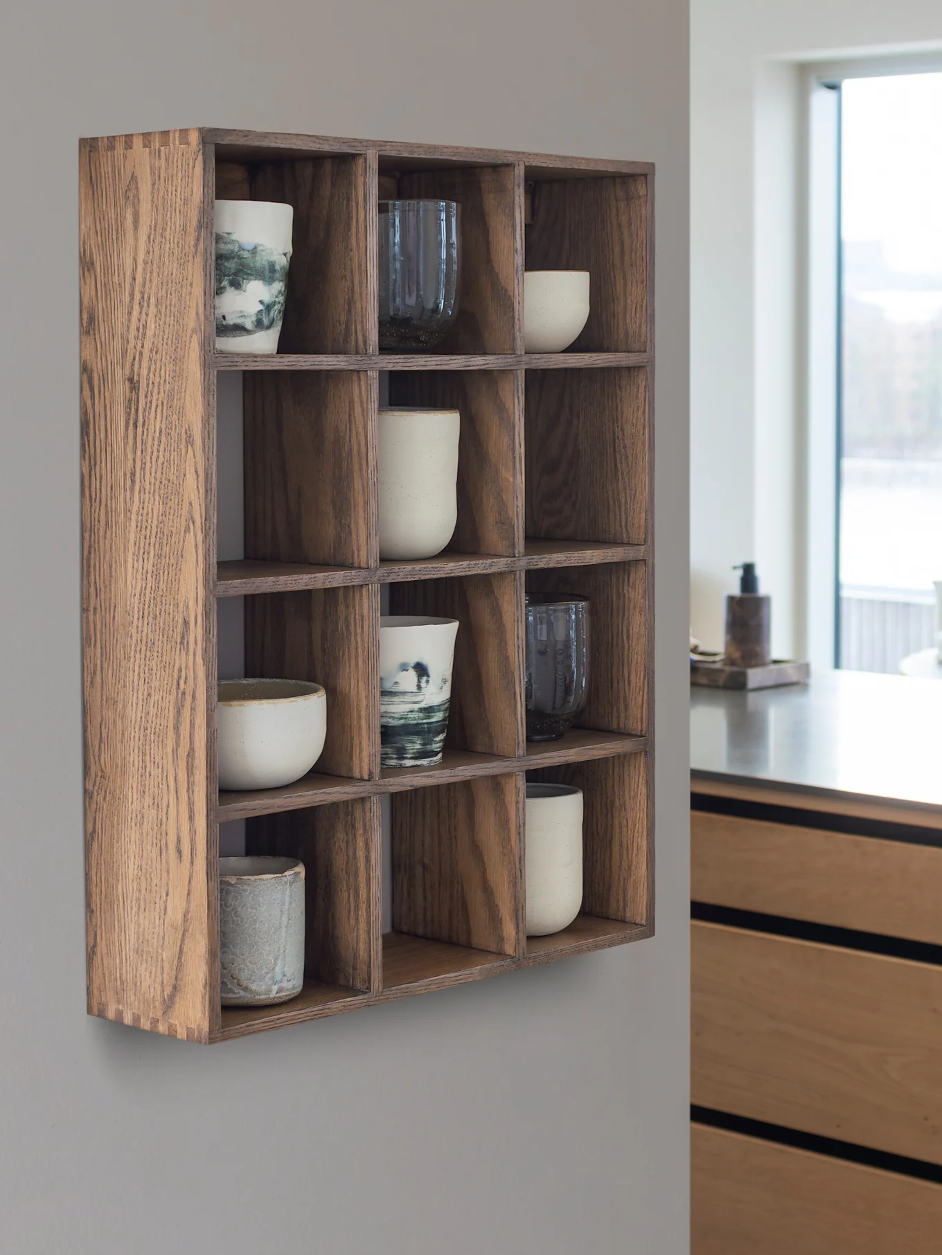 Houten wandrek met 12 vakken voor keramische kopjes en mokken. Op de achtergrond een moderne keuken en een raam.