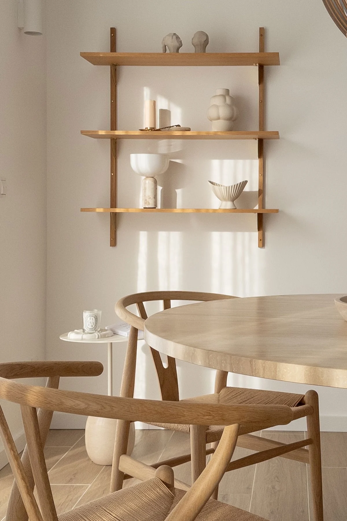 Minimalistische eetruimte met een ronde licht houten tafel en stoelen met gevlochten zittingen. Houten planken tonen decoratieve keramiek en kaarsen.