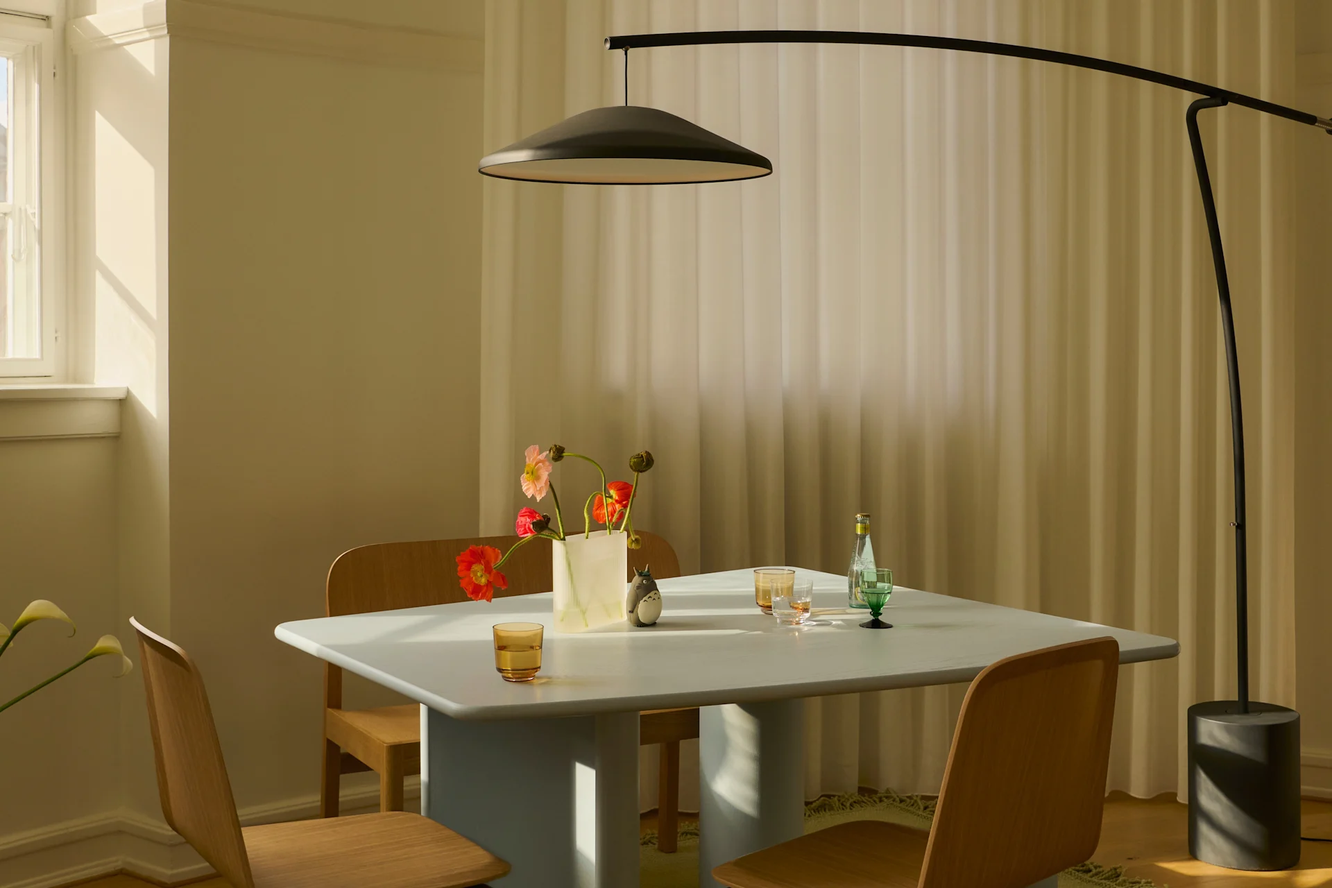 Moderne eetkamer met lichtblauwe tafel, houten stoelen, zwarte booglamp en bloemen in zonlicht.
