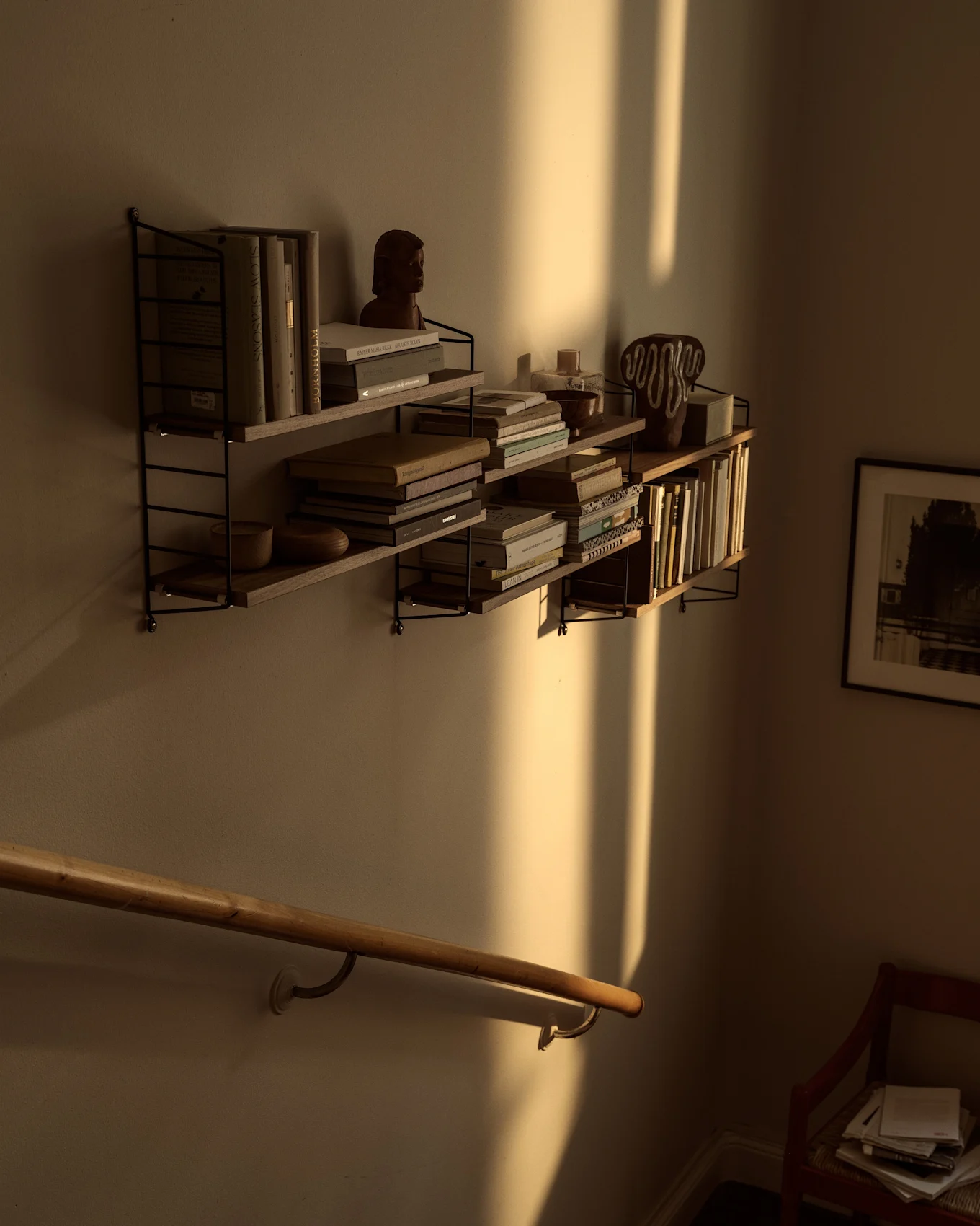 Wandplanken met boeken en decor, badend in warm zonlicht dat sterke verticale schaduwen werpt boven een houten leuning.