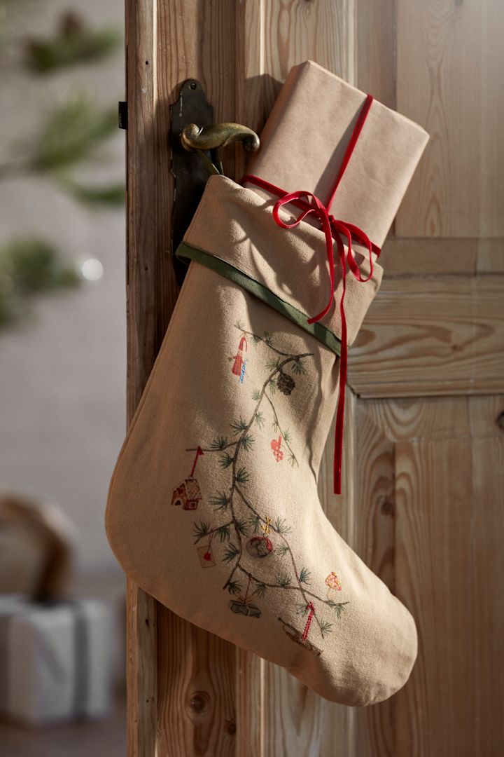 De kerstdecoraties van 2025 zijn geïnspireerd door de natuur – zoals deze kerstkous van Kähler, die je hier ziet hangen aan een houten deur, voor een kerstboom en ingepakte cadeaus.