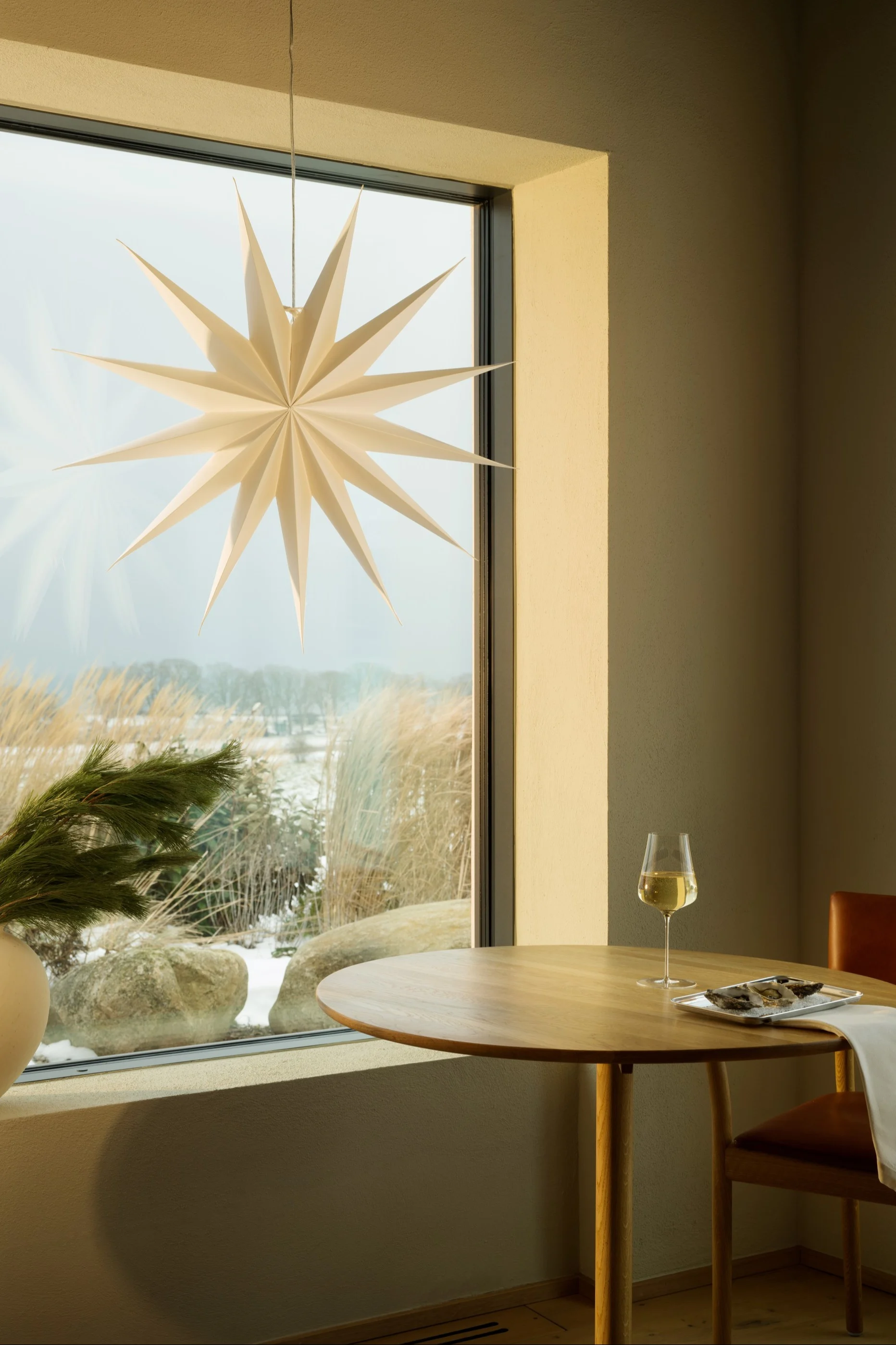 Licht venster met beige gordijnen, groenblijvende plant en witte sterlantaarn. Licht beddengoed voor.