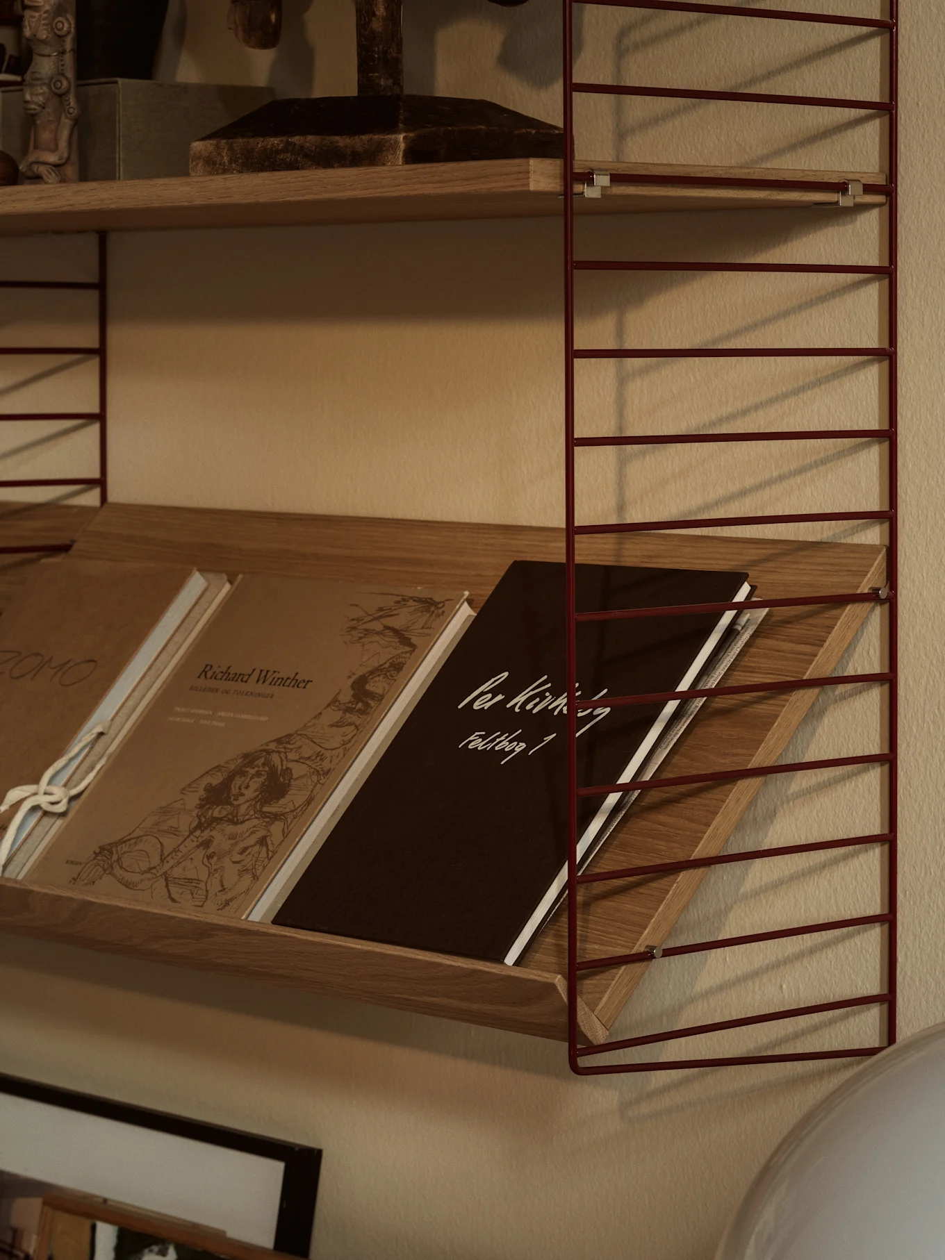 Een rood metalen draadrek met houten planken. De onderste plank bevat schuin geplaatste boeken, waaronder een donkerbruin exemplaar met 'Per Kirkeby Feldbog 1'.