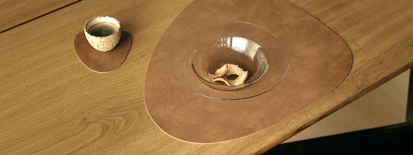 Kopje koffie op kleine bruine placemat, glazen kom met gedroogde appels op grote placemat, op houten tafel.