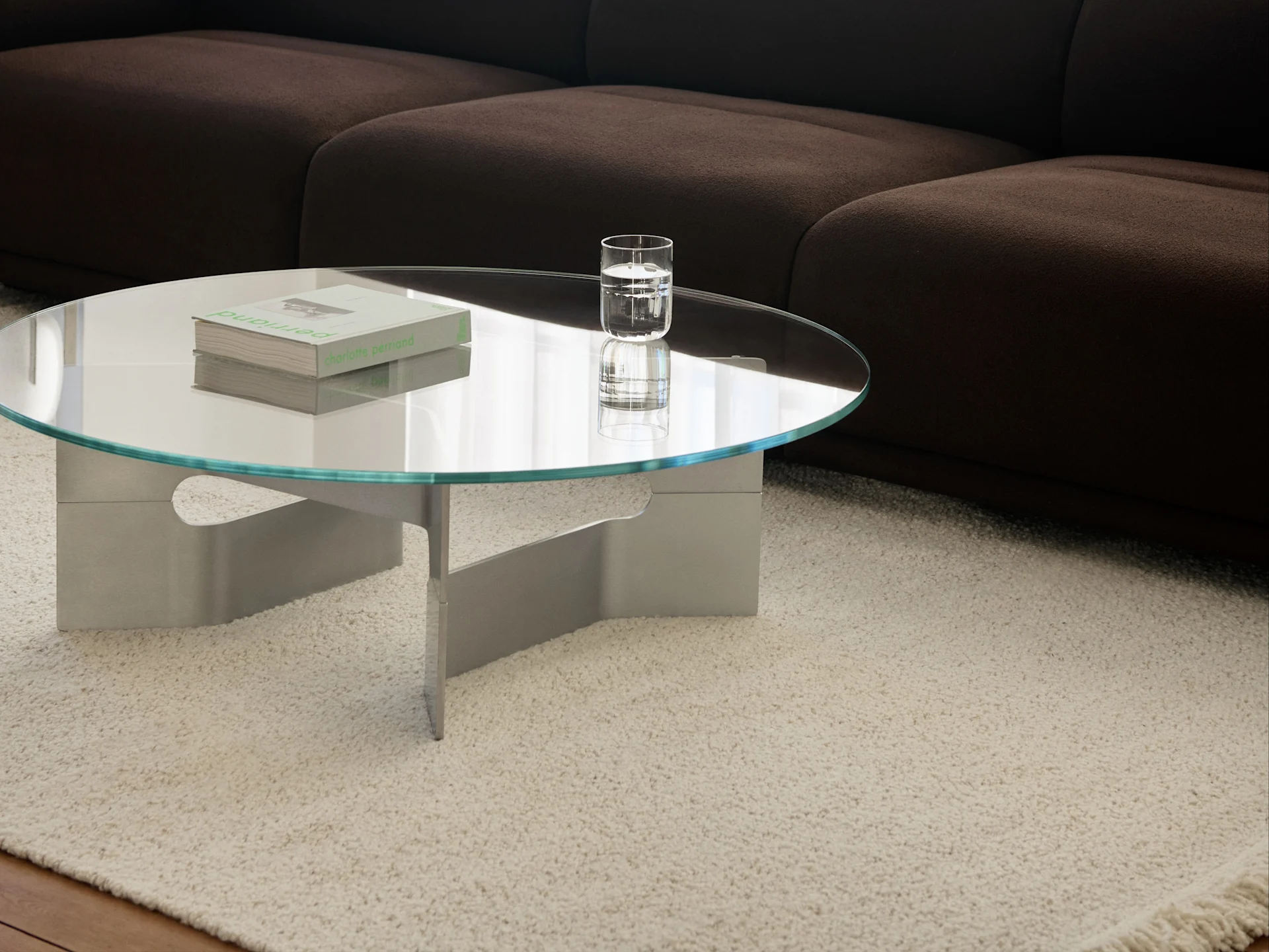 Ronde glazen salontafel met boeken en een glas water op een crèmekleurig shaggy tapijt voor een donkerbruine bank.