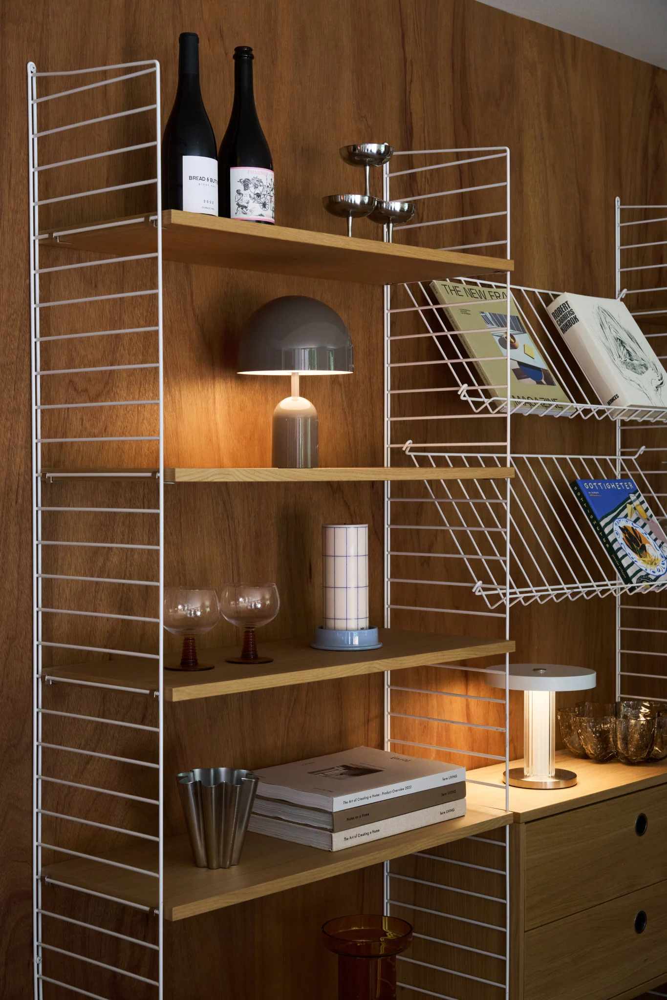Een stijlvolle houten wandplank met witte draadframes bevat boeken, flessen, een paddestoellamp en diverse decoratieve items.