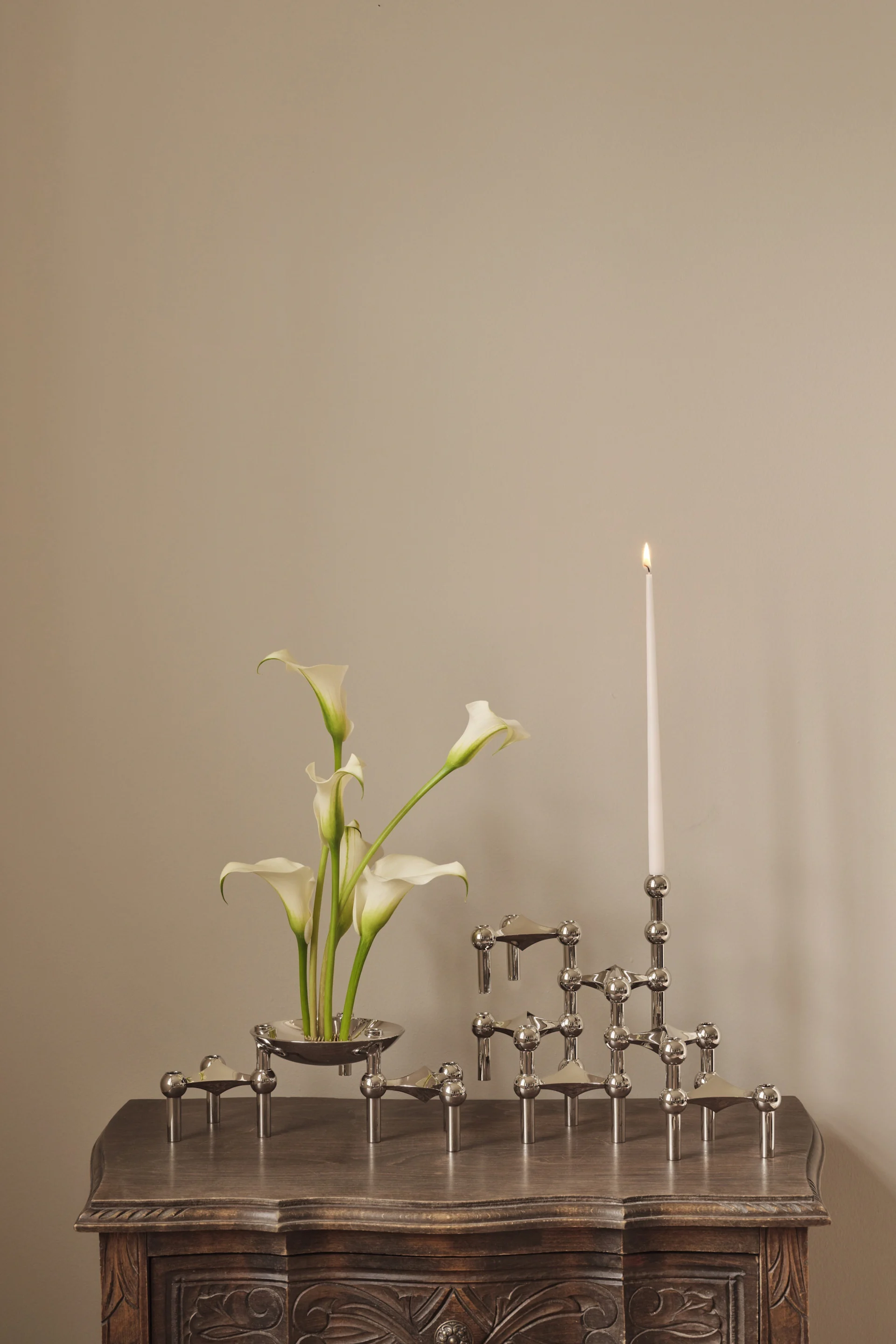 Witte calla lelies in een zilveren schaal naast een modulaire zilveren kandelaar met één brandende witte kaars op een gesneden houten tafel.