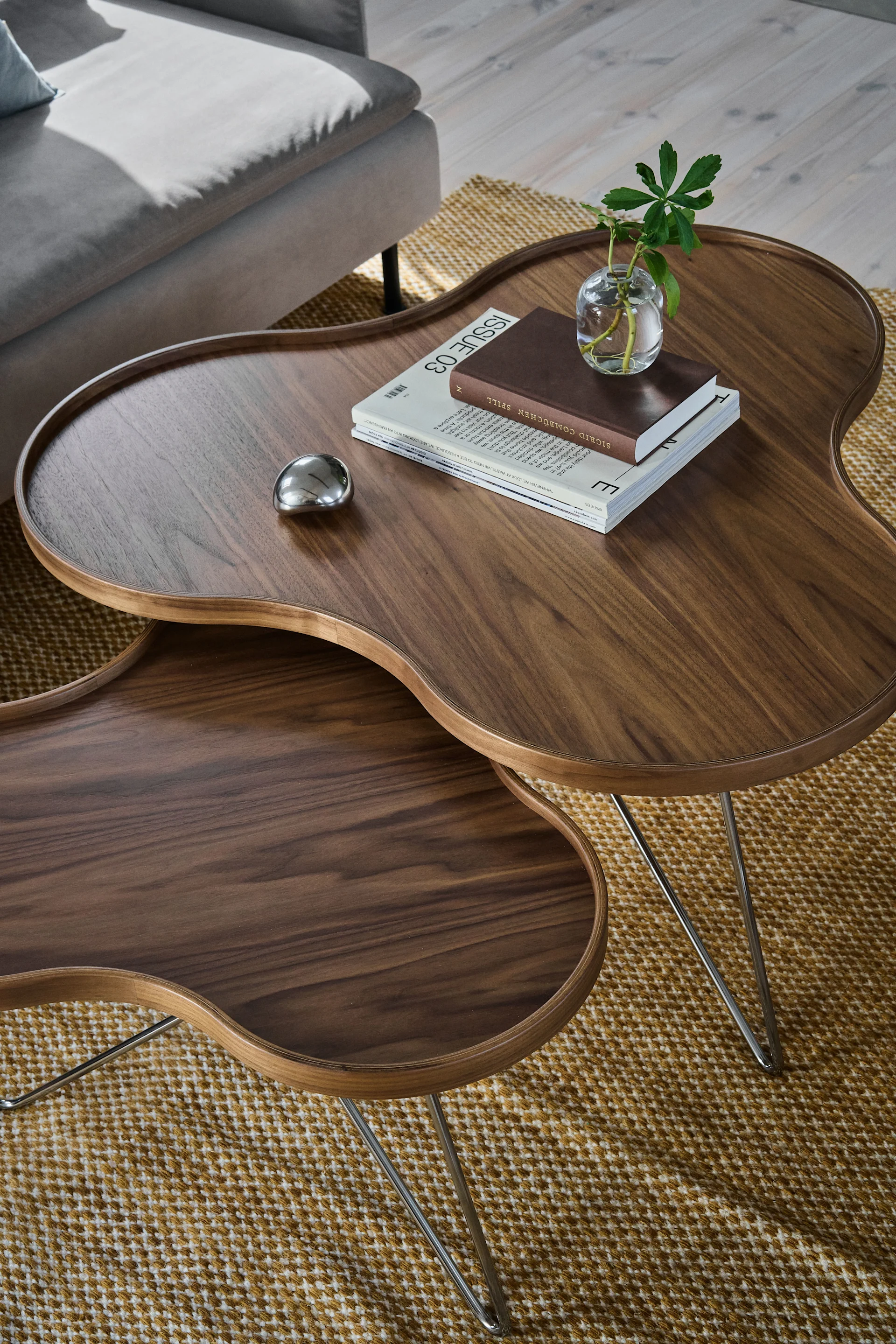 Twee donkere houten, niervormige nestbare salontafels op een gemêleerd vloerkleed, met een plant, boeken en een metalen object.
