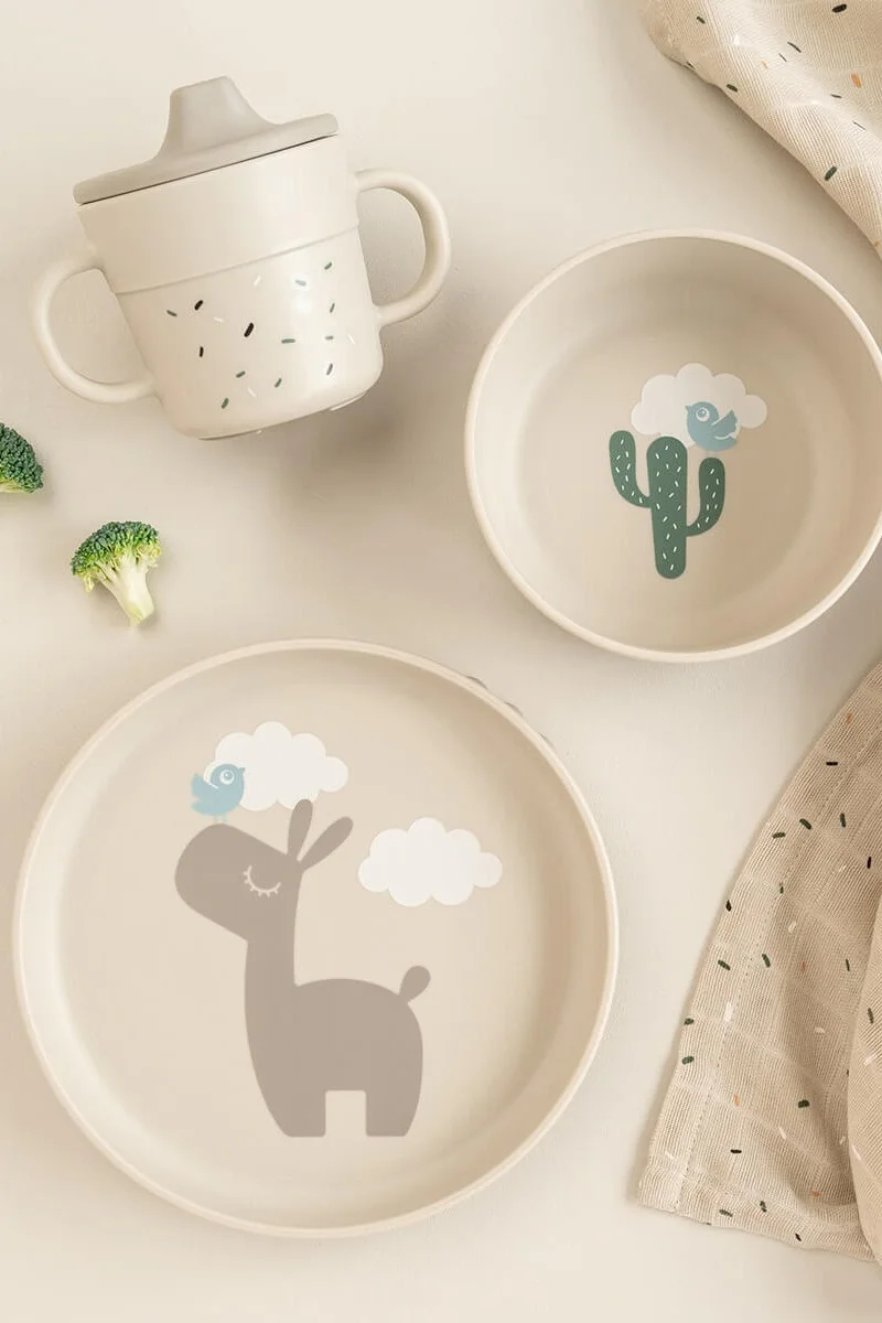 Een lichtbeige kinderservies set met een tuitbeker, een kom met een cactus en een bord met een lama en vogels.