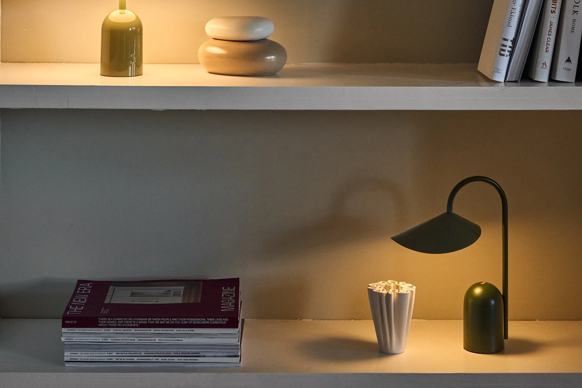 Drie planken tonen diverse tafellampen in groen en paars, samen met boeken, tijdschriften en decoratieve objecten.