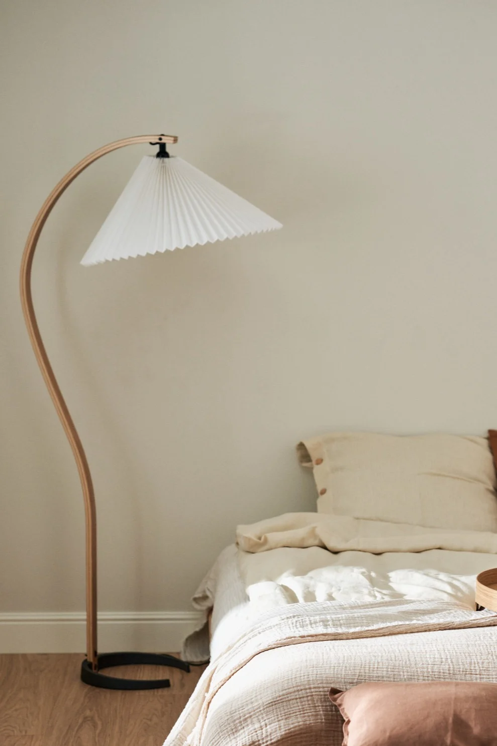 Hier zie je de Gubi Timberline vloerlamp met zijn gebogen houten poot naast een bed staan. 