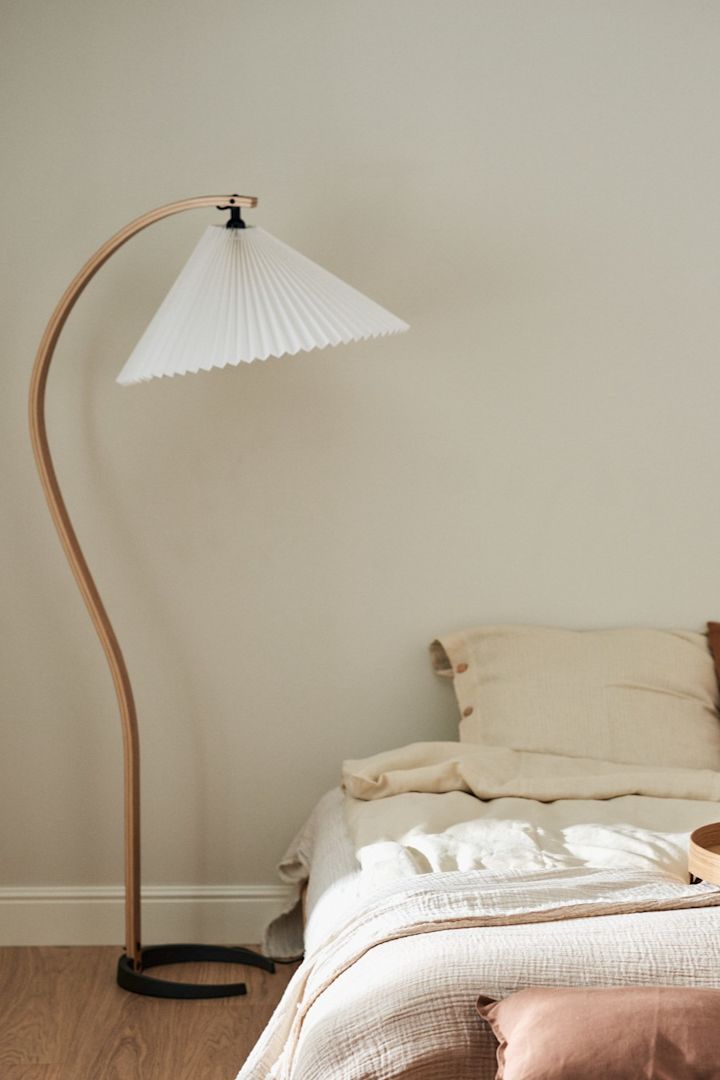 Hier zie je de Gubi Timberline vloerlamp met zijn gebogen houten poot naast een bed staan. 
