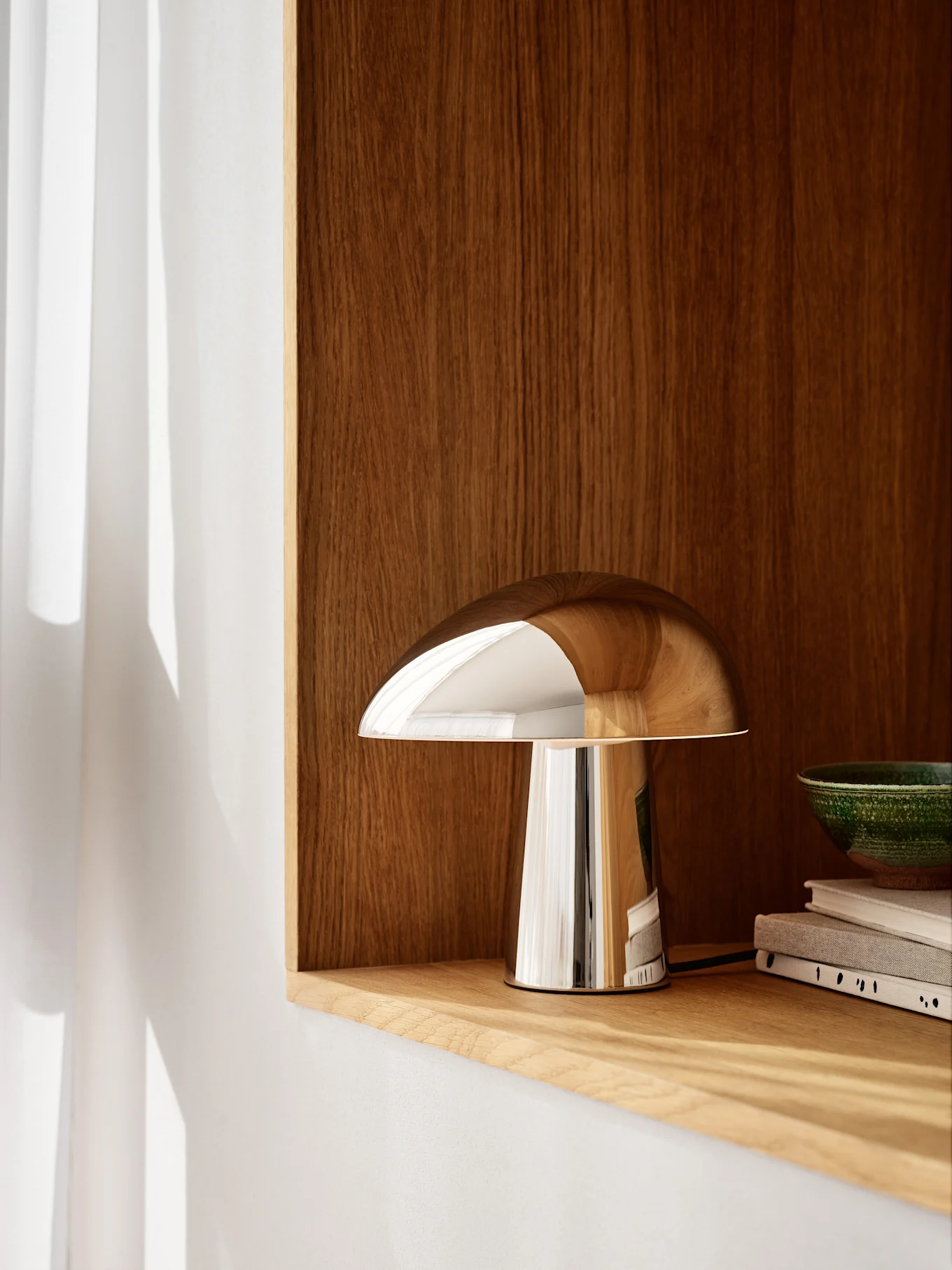 Een gepolijste chromen paddestoelvormige lamp staat op een lichte houten plank, met een donkere houten wand erachter. Een groene kom en boeken staan rechts.