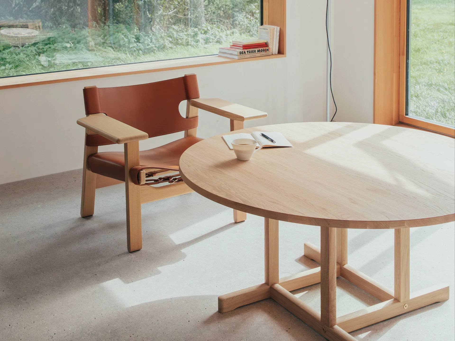 Lichte kamer met bruine leren fauteuil, ronde houten tafel met kopje en notitieboekje, zonovergoten door raam met uitzicht op groen.