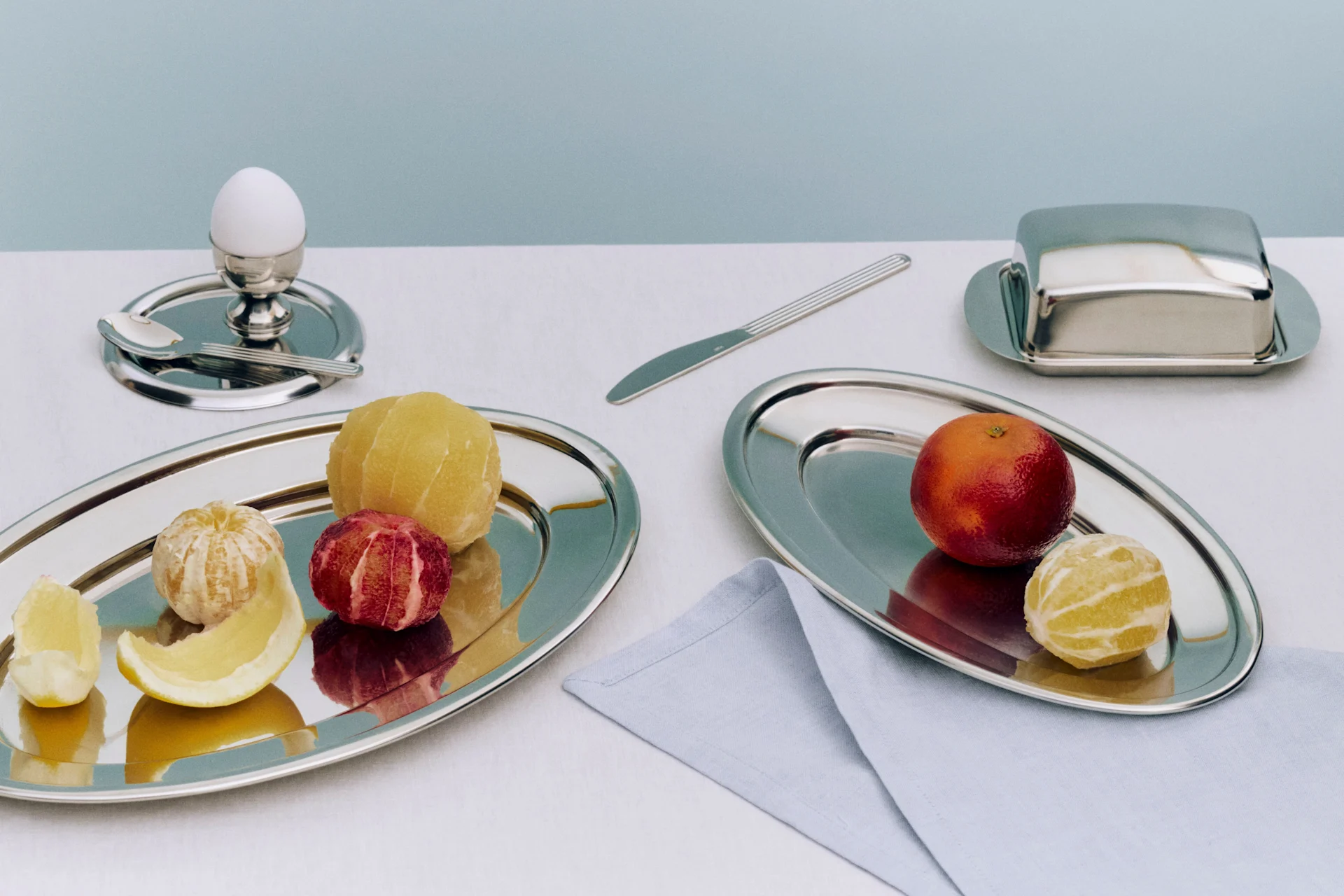 Ontbijt met geschilde en ongeschilde citrusvruchten op zilveren schalen, een ei in eierdop en een botervloot.