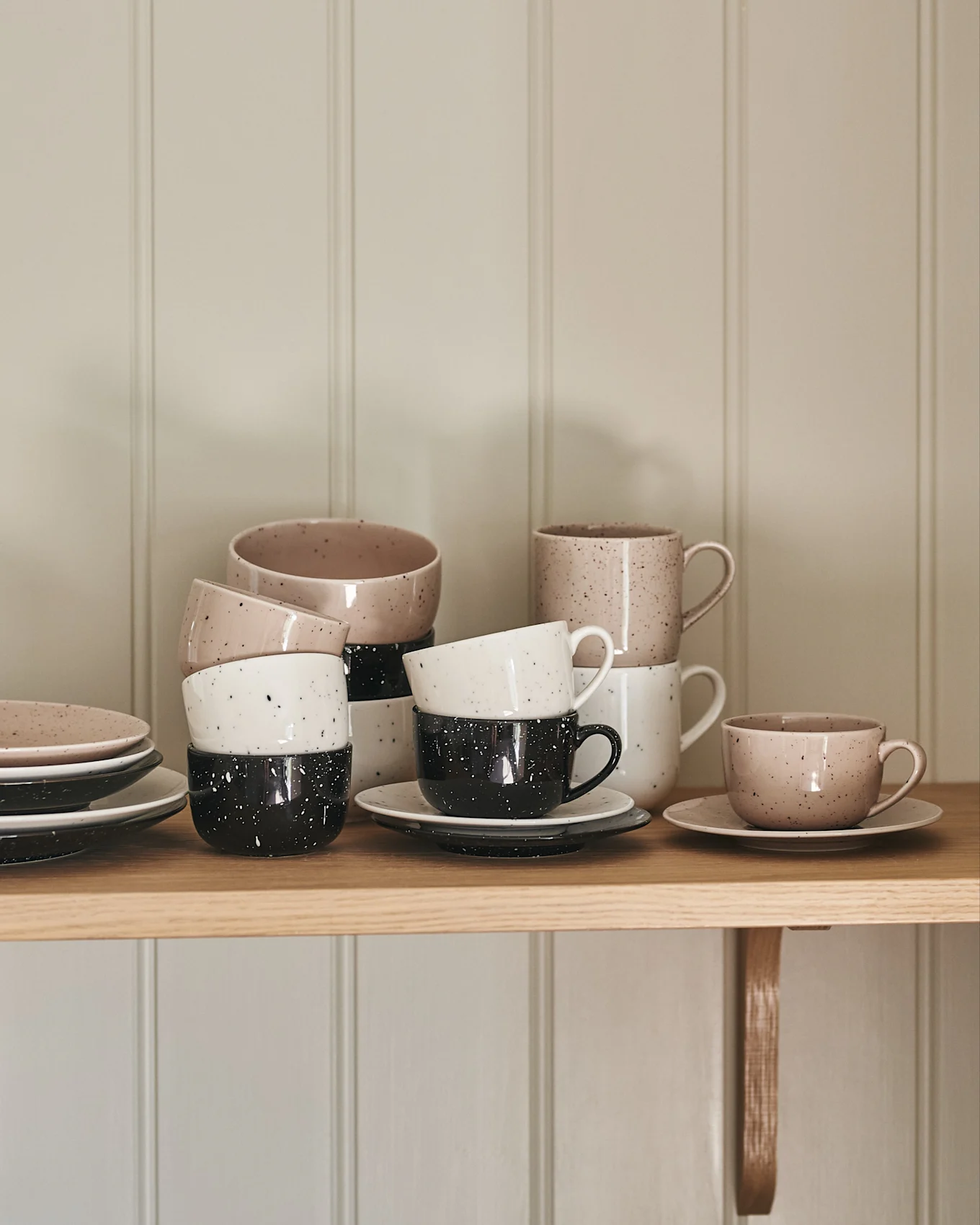 Gestapelde keramische kopjes en borden in gespikkeld beige, wit en zwart op een houten plank.