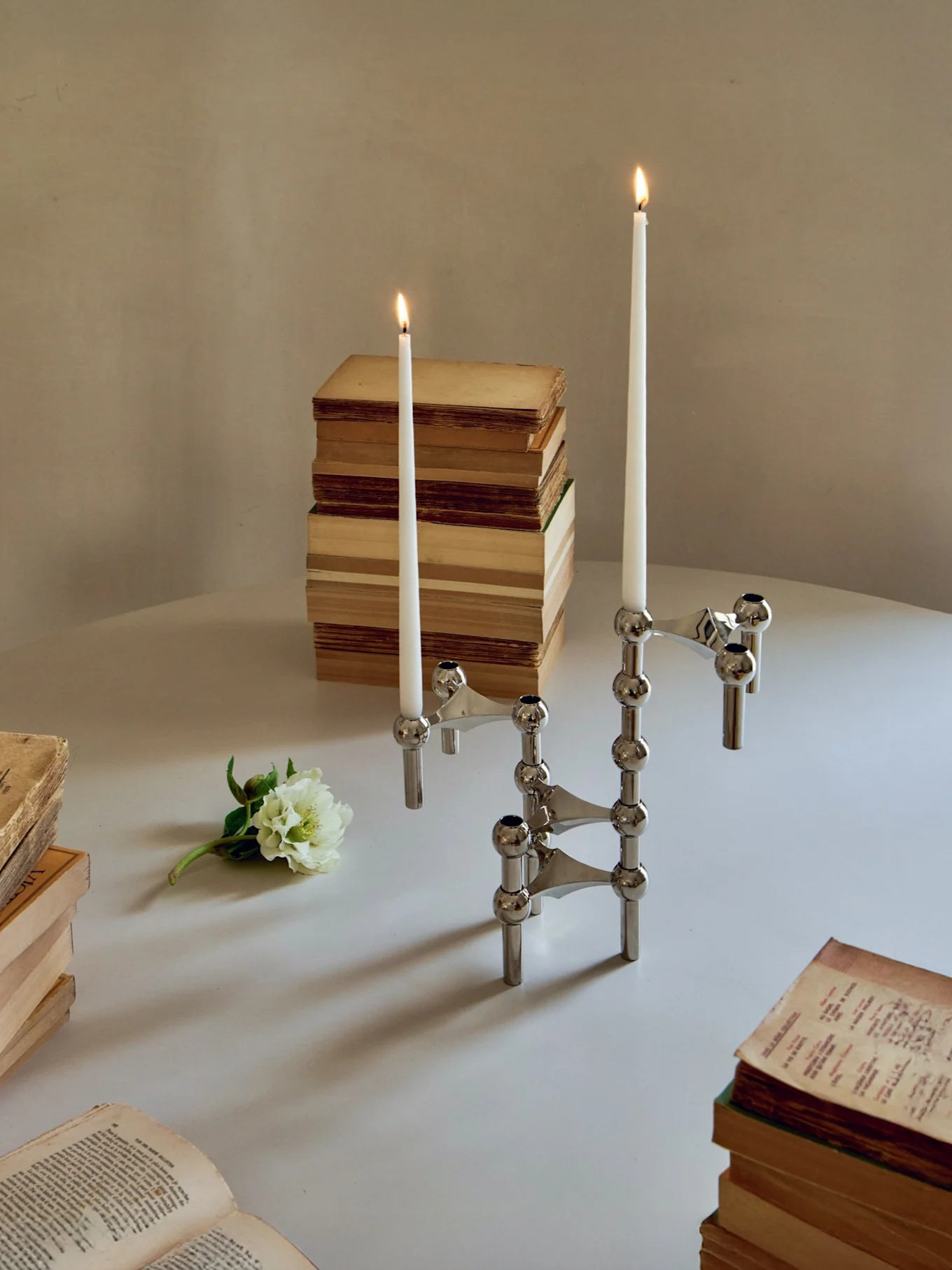 Een zilveren modulaire kandelaar met twee brandende kaarsen, boeken en een witte bloem op een tafel.