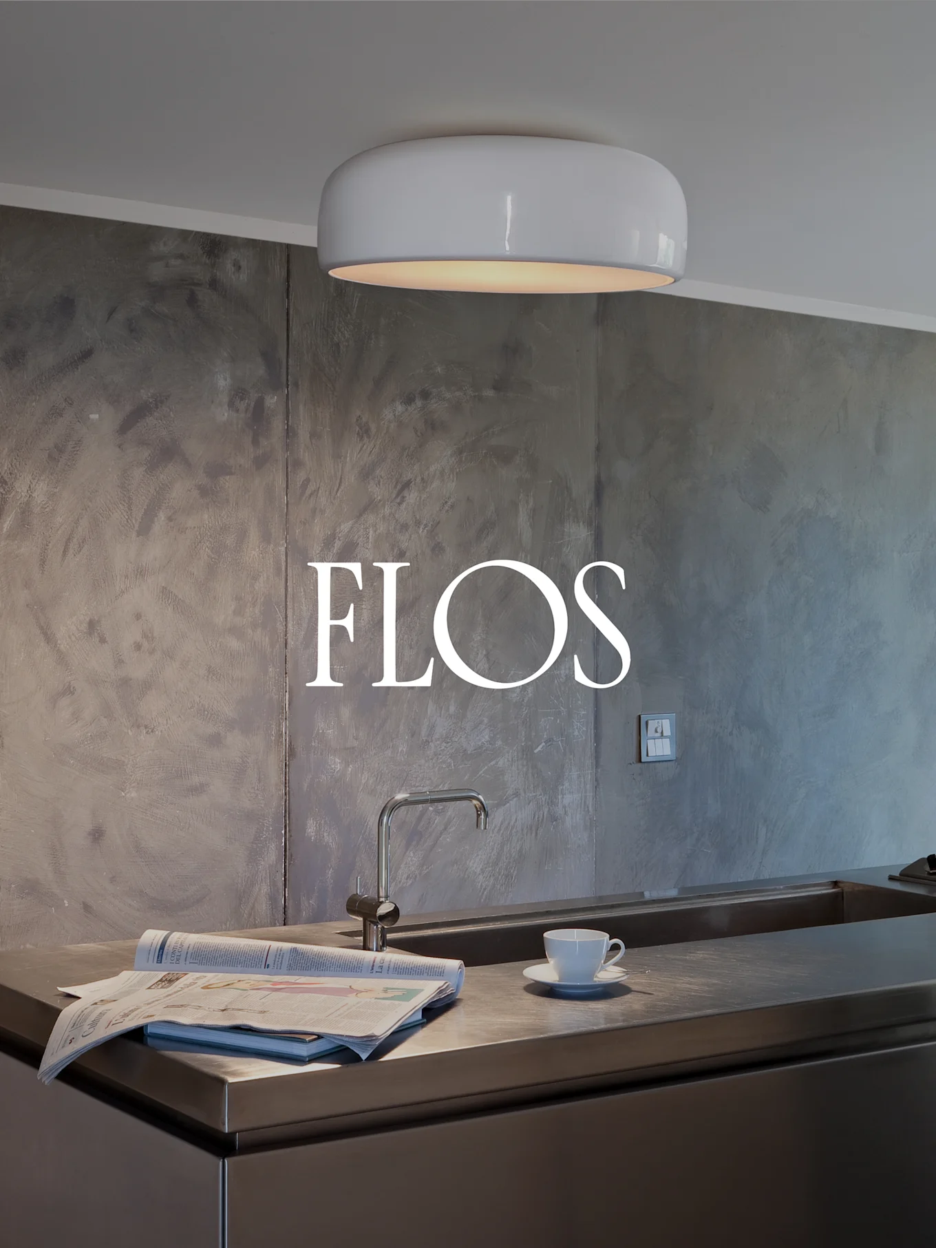 Moderne keuken met witte, afgeronde plafondlamp, roestvrijstalen aanrecht met spoelbak, krant en theekopje. FLOS-logo op gestructureerde muur.