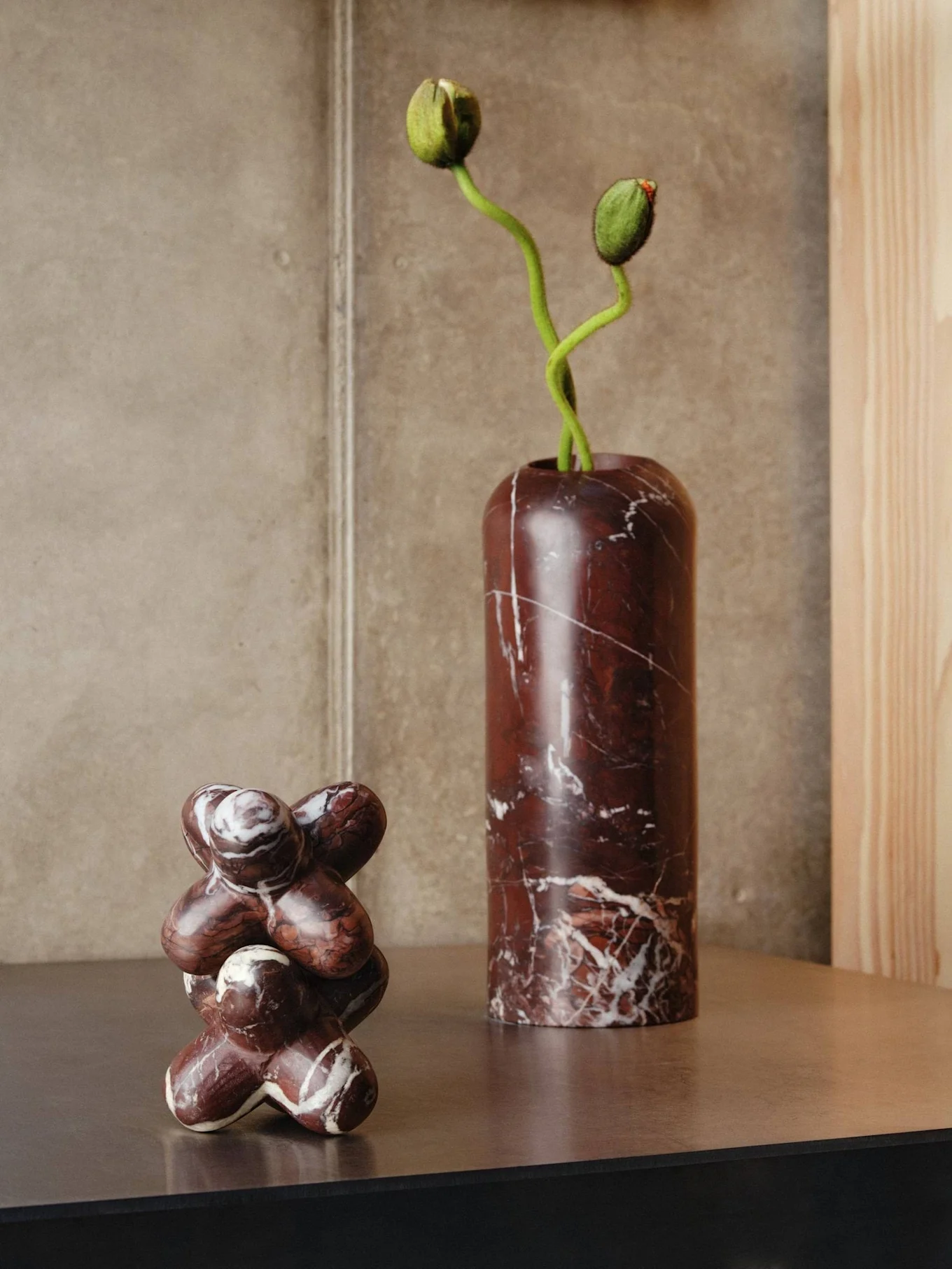 Twee marmeren objecten op een plank: een hoge donkerrode vaas met groene bloemknoppen en een abstract sculptuur.