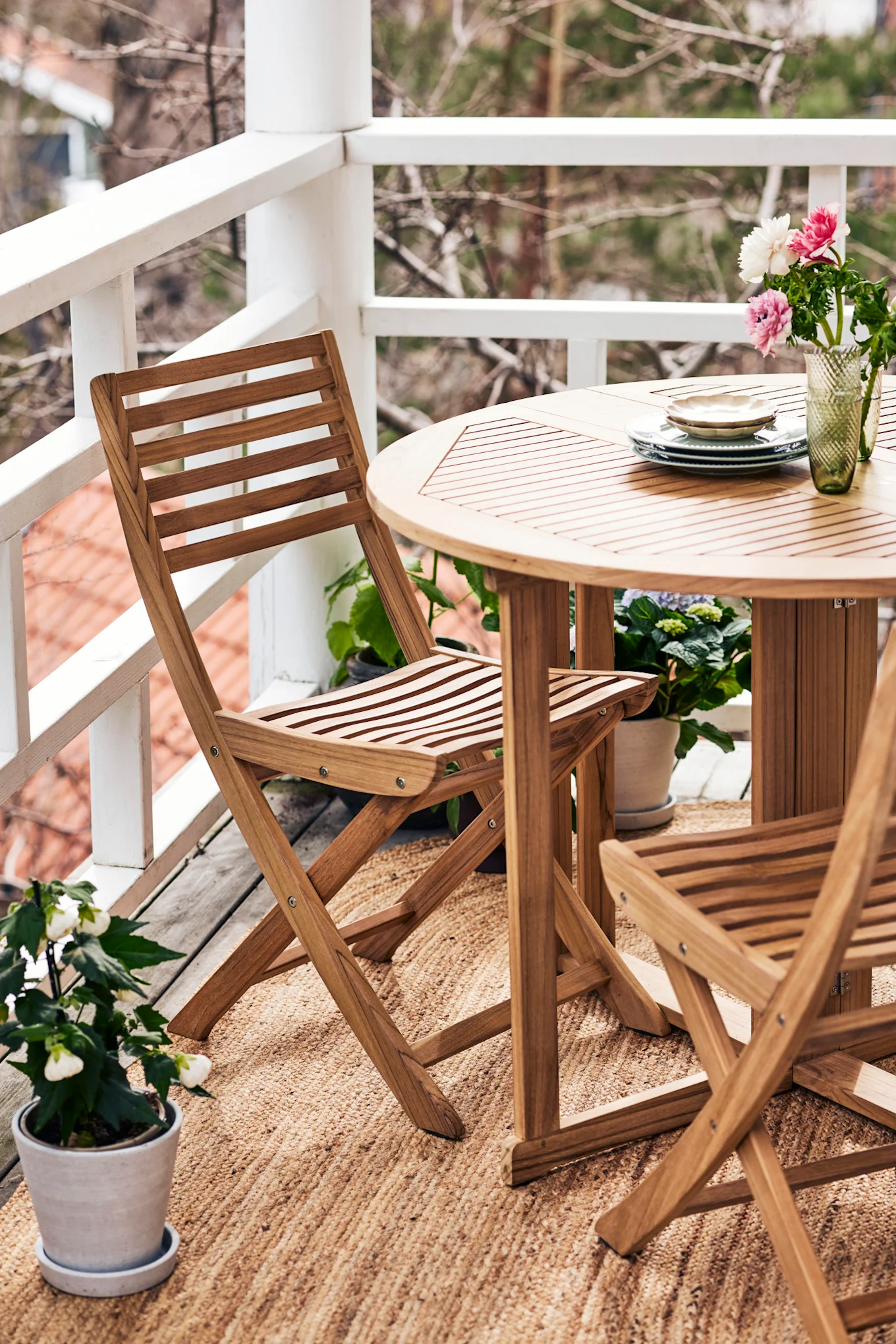Balkon met ronde lattenhouten tafel, twee klapstoelen op geweven vloerkleed, vaas bloemen en gestapelde borden.