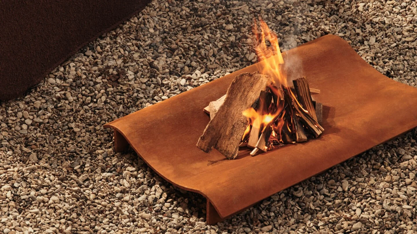 Klein kampvuur met brandend hout op een rechthoekig, roestig metalen dienblad op grind.