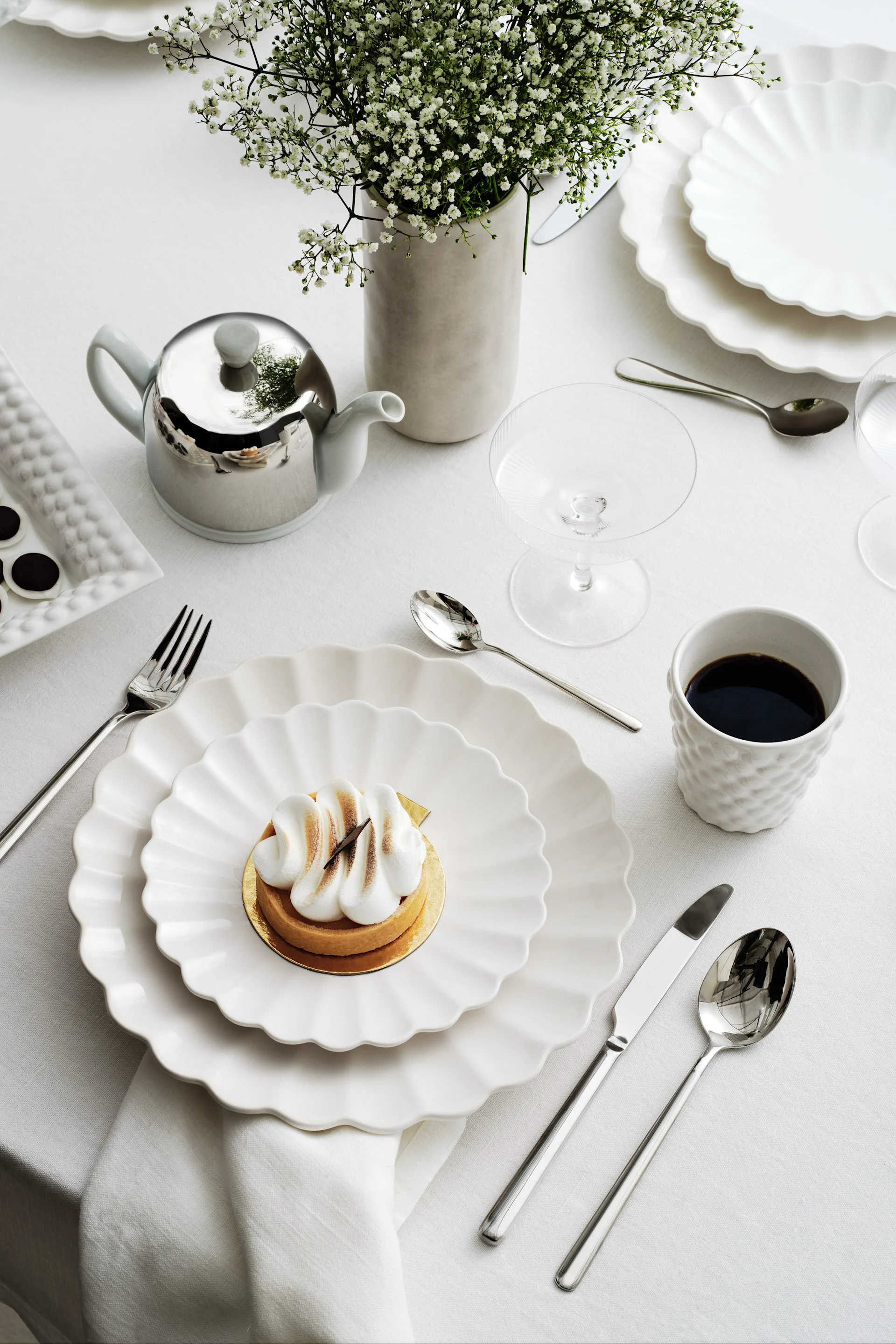Elegante tafeldekking met witte geschulpte borden, een merengue taartje, koffie, een zilveren theepot en gipskruid.