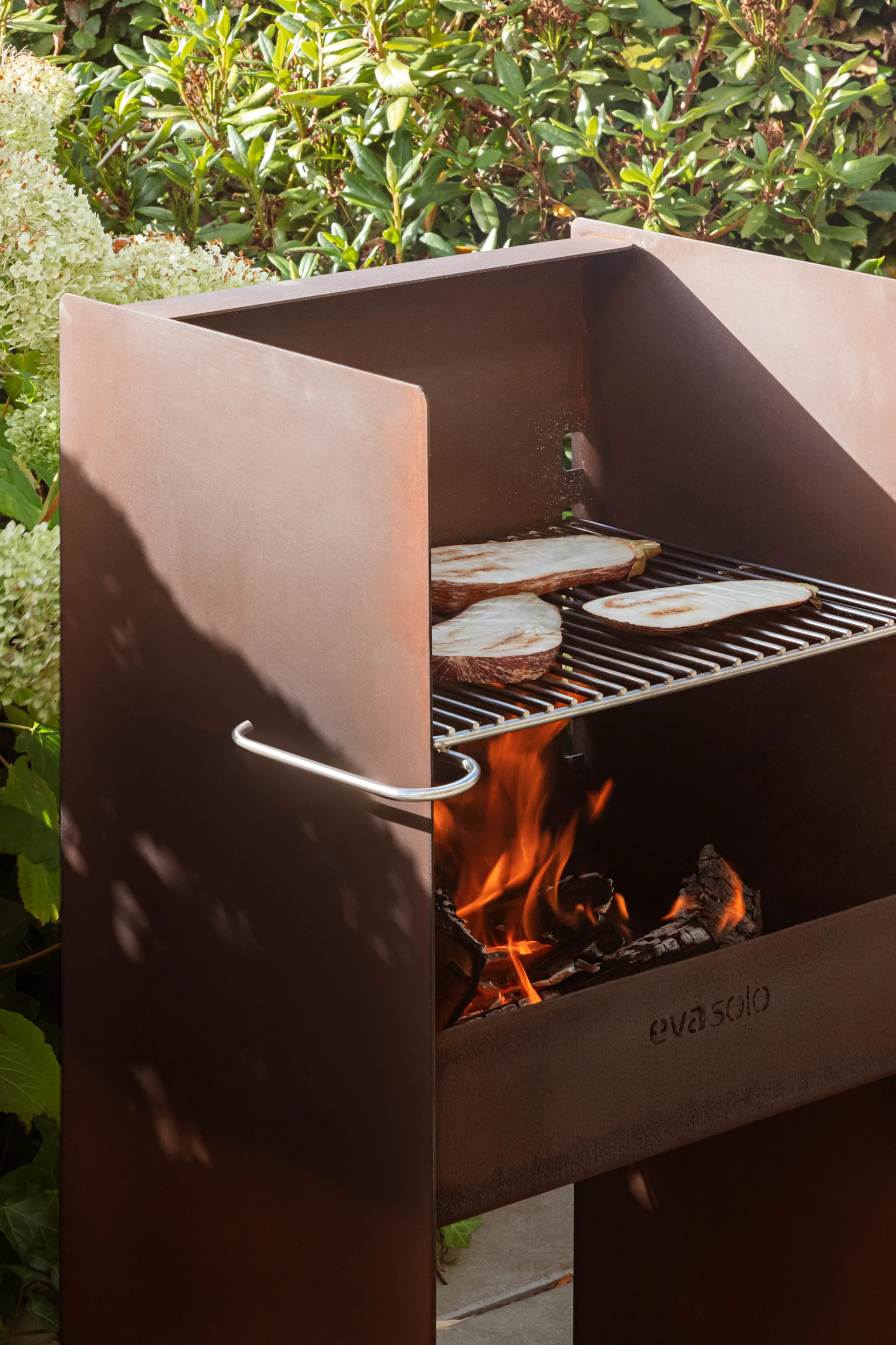 Cortenstaal buitenbarbecue met aubergineplakken die op een rooster boven open vuur worden bereid, omringd door groene planten.