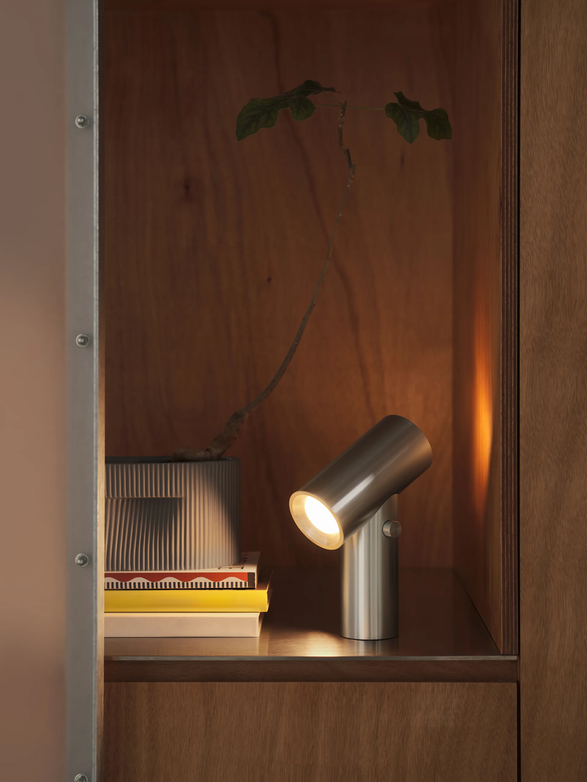 Een geborstelde metalen cilindrische lamp verlicht een stapel boeken en een plant in een geribbelde pot, in een houten kast.