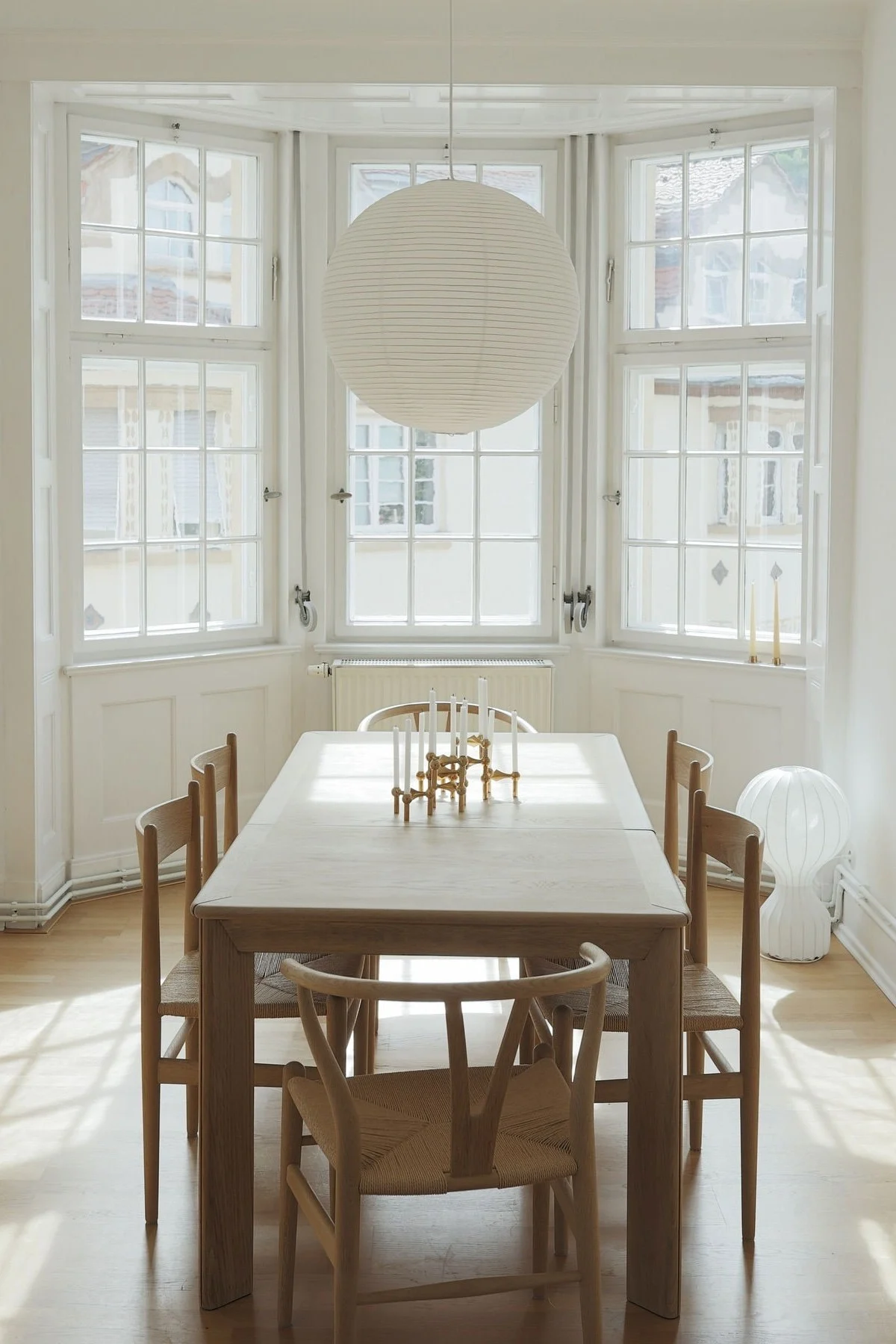 Lichte eetkamer met een licht houten tafel, zes stoelen, een grote papieren lantaarn en een gouden kandelaar op tafel. Zonlicht stroomt door de erkers.