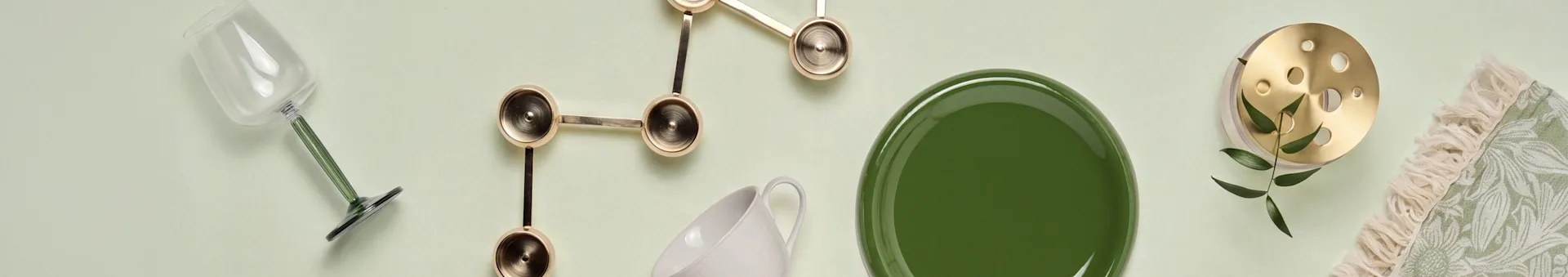 Glas met groene steel, gouden sculpturaal decor, witte mok, groen bord, messing item met bladeren, groen textiel met franjes.