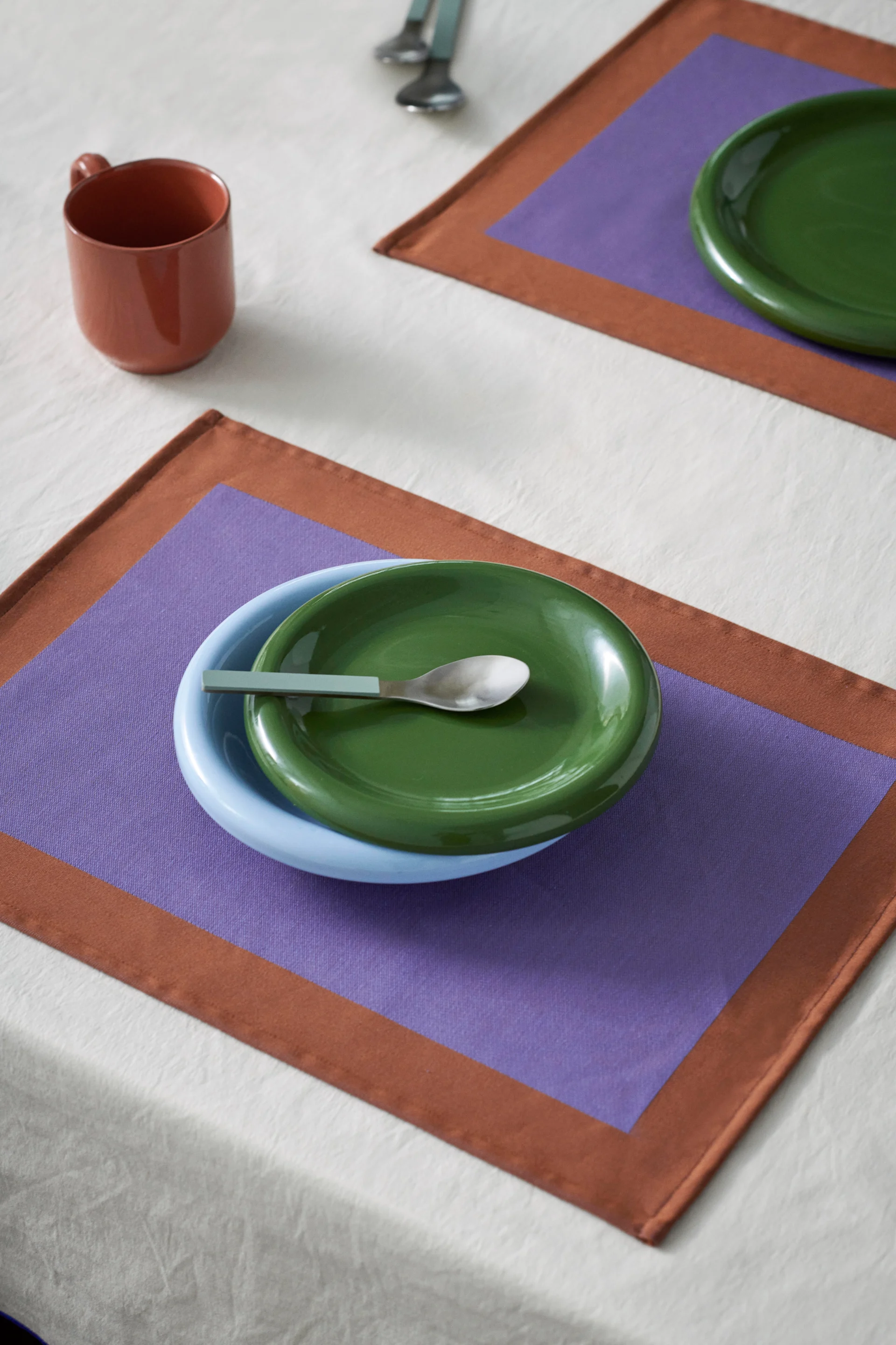 Gedekte tafel met blauwe kom, groen bord en lepel op paars/bruin placemat. Een bruine mok staat op het witte tafelkleed.