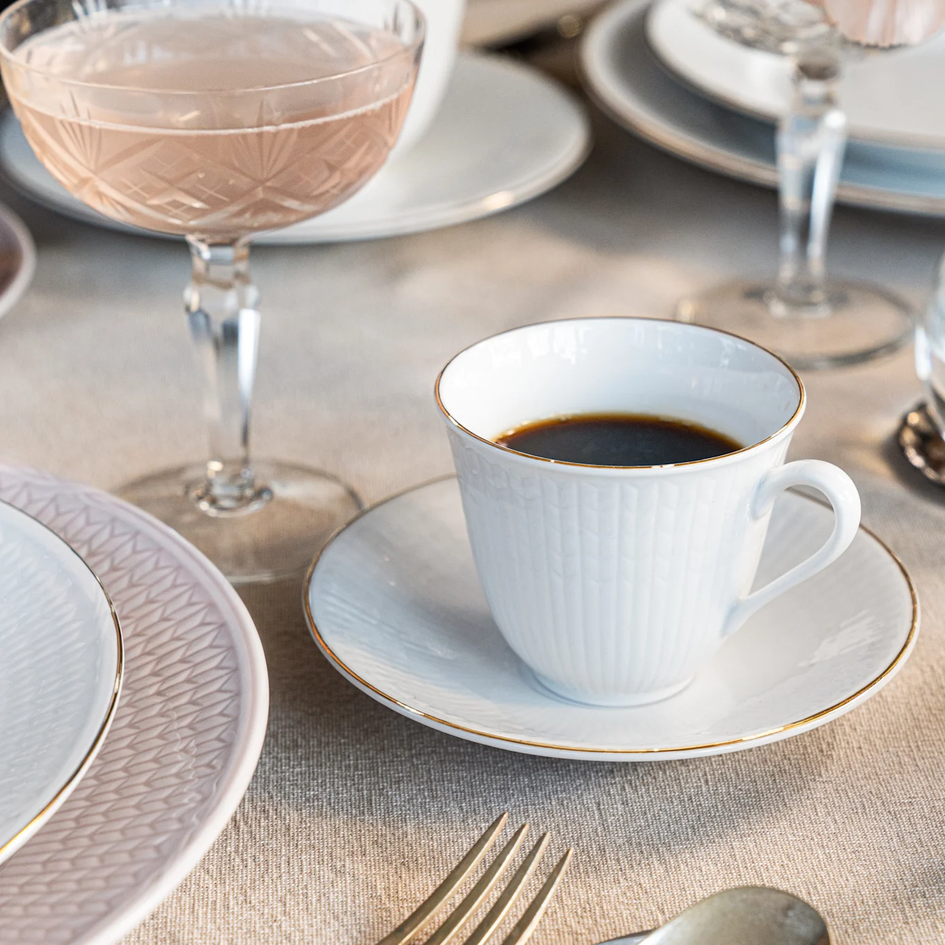 Een witte koffiekop met gouden rand op een schotel, naast een roze bord en een roséglas op een linnen tafelkleed.