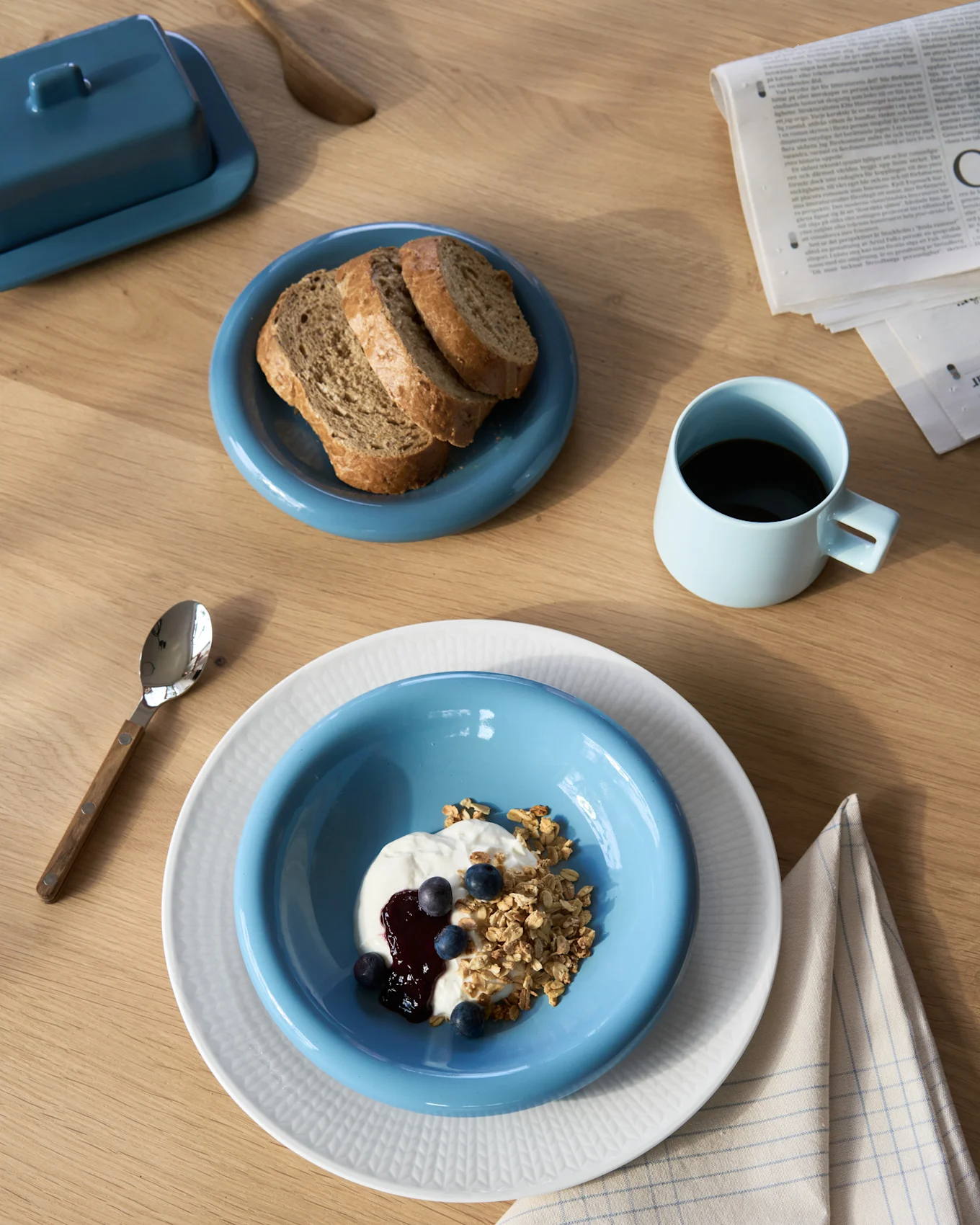 Bovenaanzicht van een ontbijt met yoghurt en granola, donker brood, koffie en een botervloot op een houten tafel.