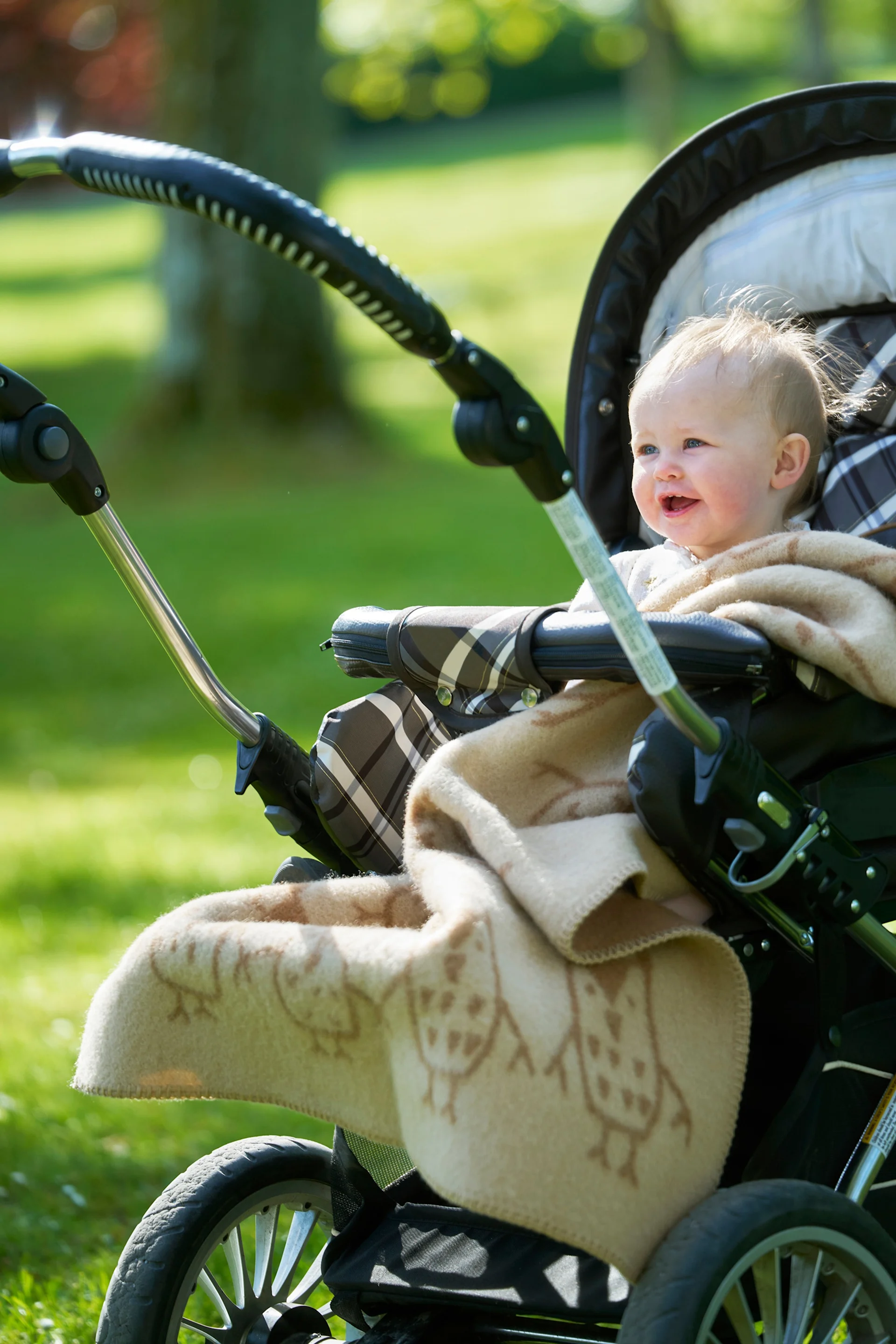 Een lachende baby met licht haar, gewikkeld in een deken met uilenpatroon, zit buiten in een kinderwagen in een zonnig park.