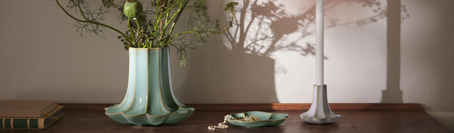 Een geribbelde teal vaas met bladeren, een parelketting in een bijpassend schaaltje, een witte kaars in een geribbelde houder en boeken op een houten oppervlak.