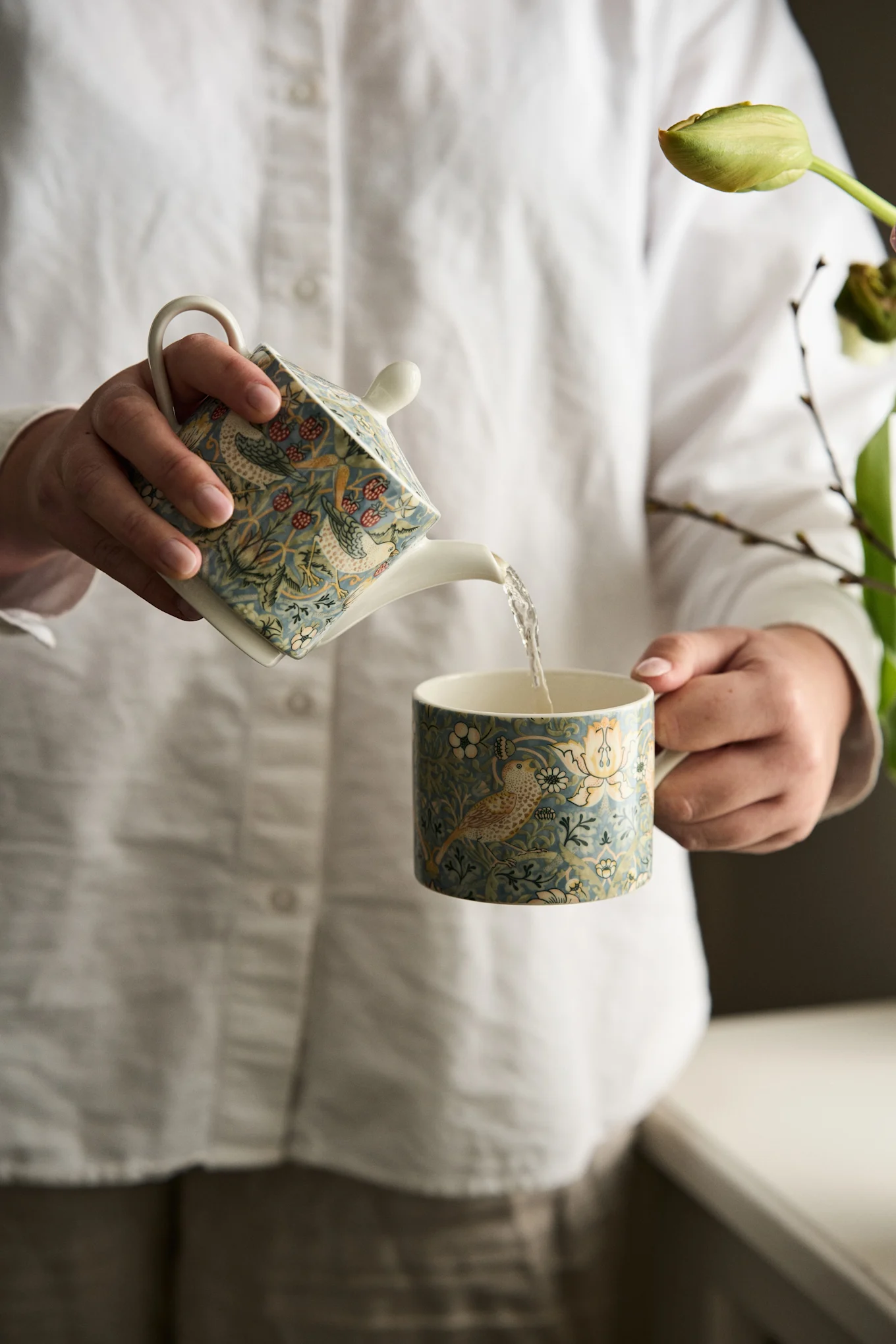 Handen schenken uit een blauwe theepot met vogel- en bessenpatroon in een bijpassende mok.