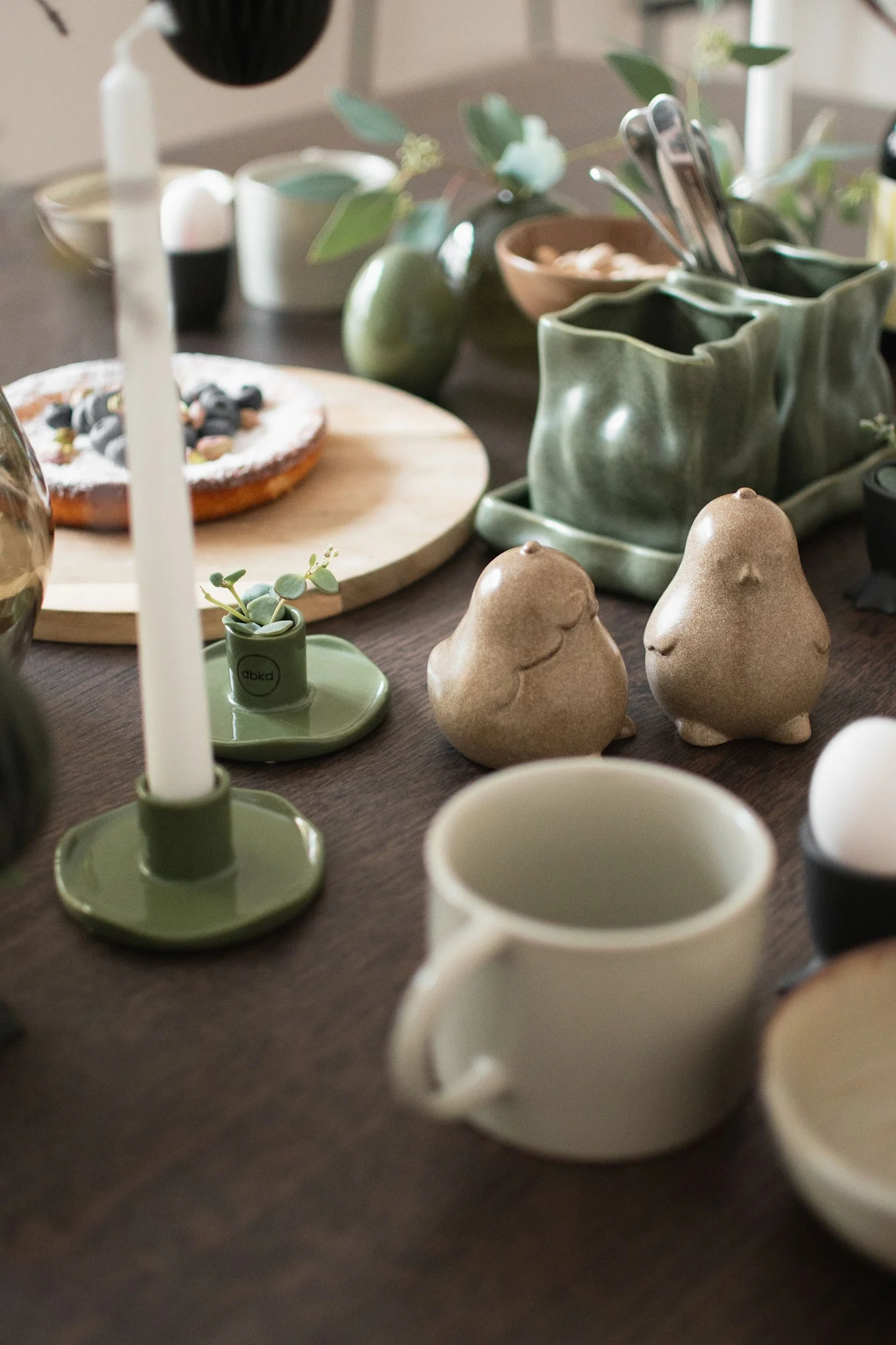 Tafel gedekt met groen keramiek servies, een blauwbessentaart, kaarsen en twee vogelvormige shakers.