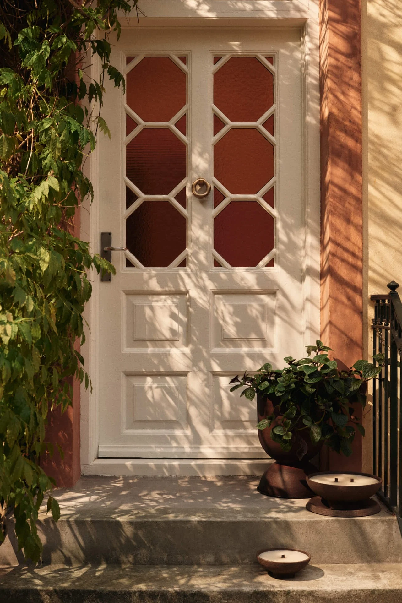 Witte voordeur met achthoekige glaspanelen, groene klimop aan de linkerkant en een plantenbak met twee kaarsen op de treden.