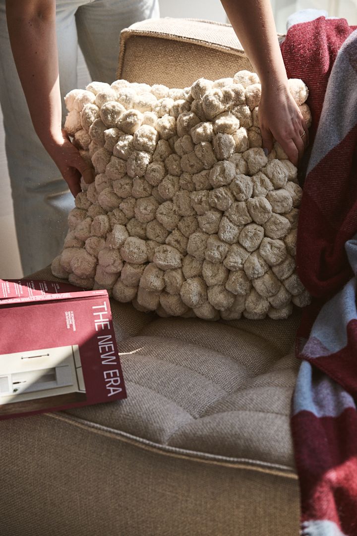 Een ander zeer geliefd product van Byon is het Pom-pom kussen, hier in offwhite op een beige bank met een gestreepte dekentje in wijnrood en lichtblauw ernaast.