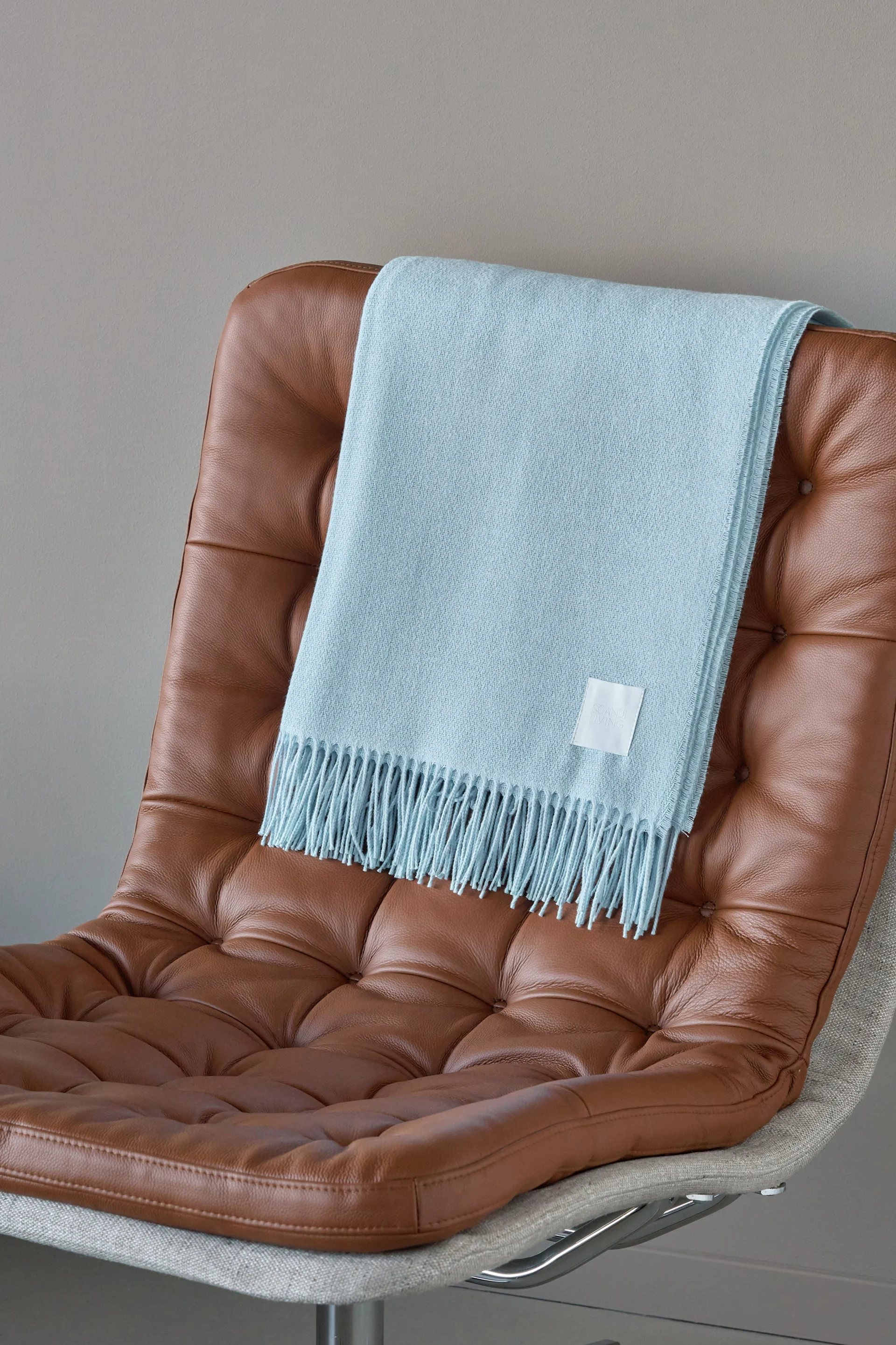 Lichtblauwe deken met franjes over een bruine, getufte leren fauteuil gedrapeerd.