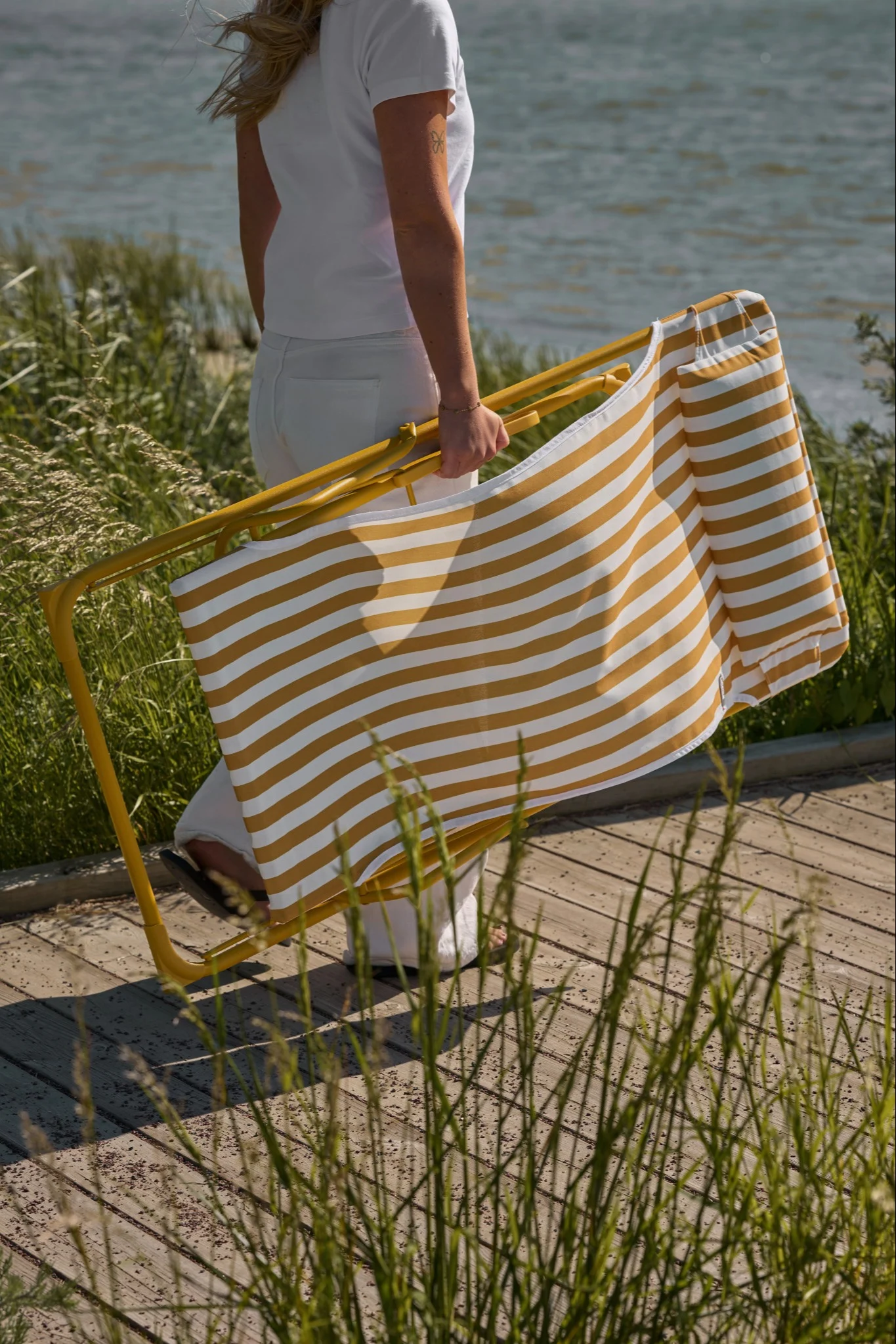 Persoon in wit draagt een opgevouwen geel-wit gestreepte strandstoel op een vlonder met hoog gras bij het water.