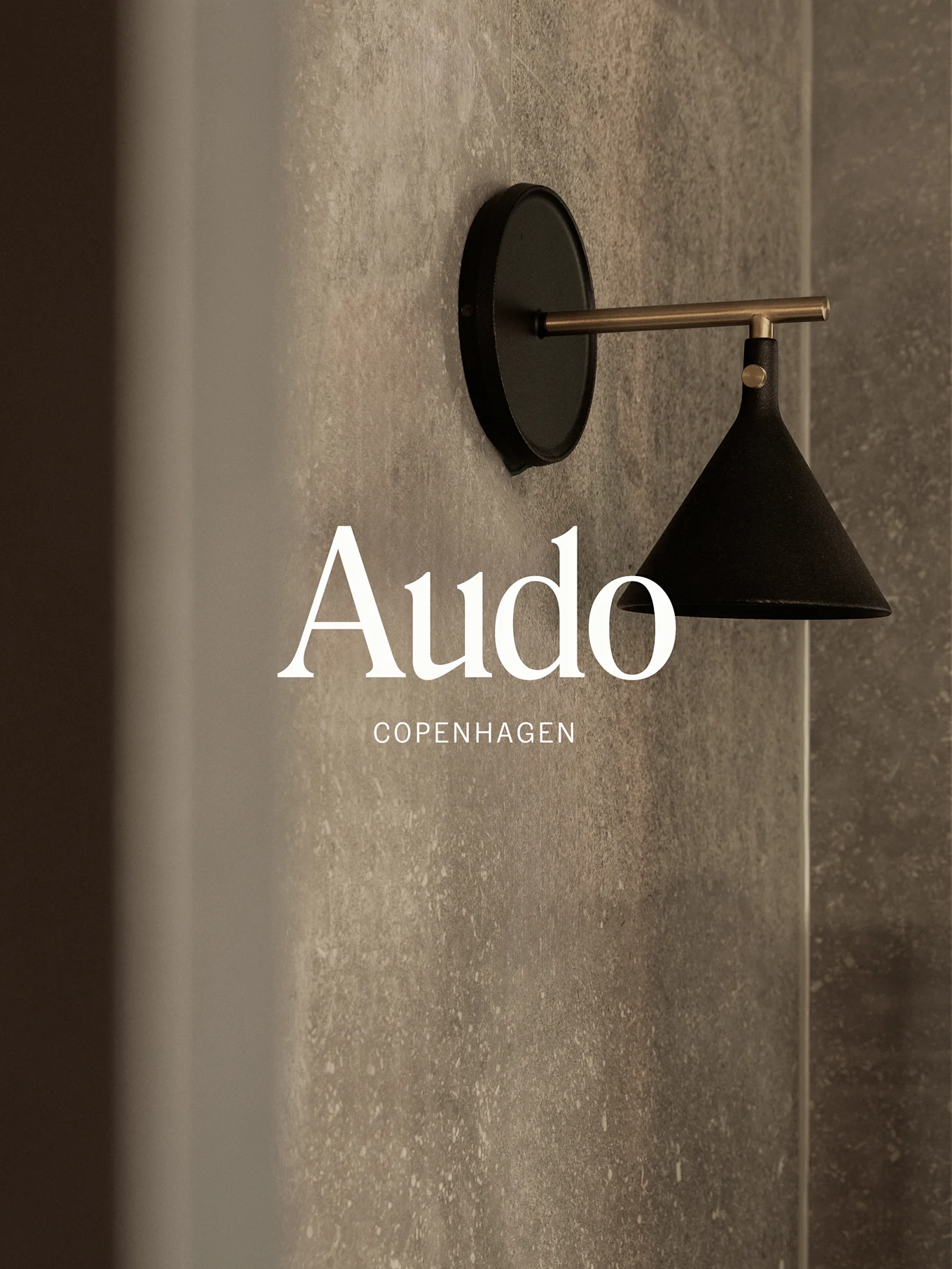 Een wandgemonteerde zwarte lamp met een messing arm tegen een getextureerde grijze muur, met de tekst 'Audo Copenhagen'.